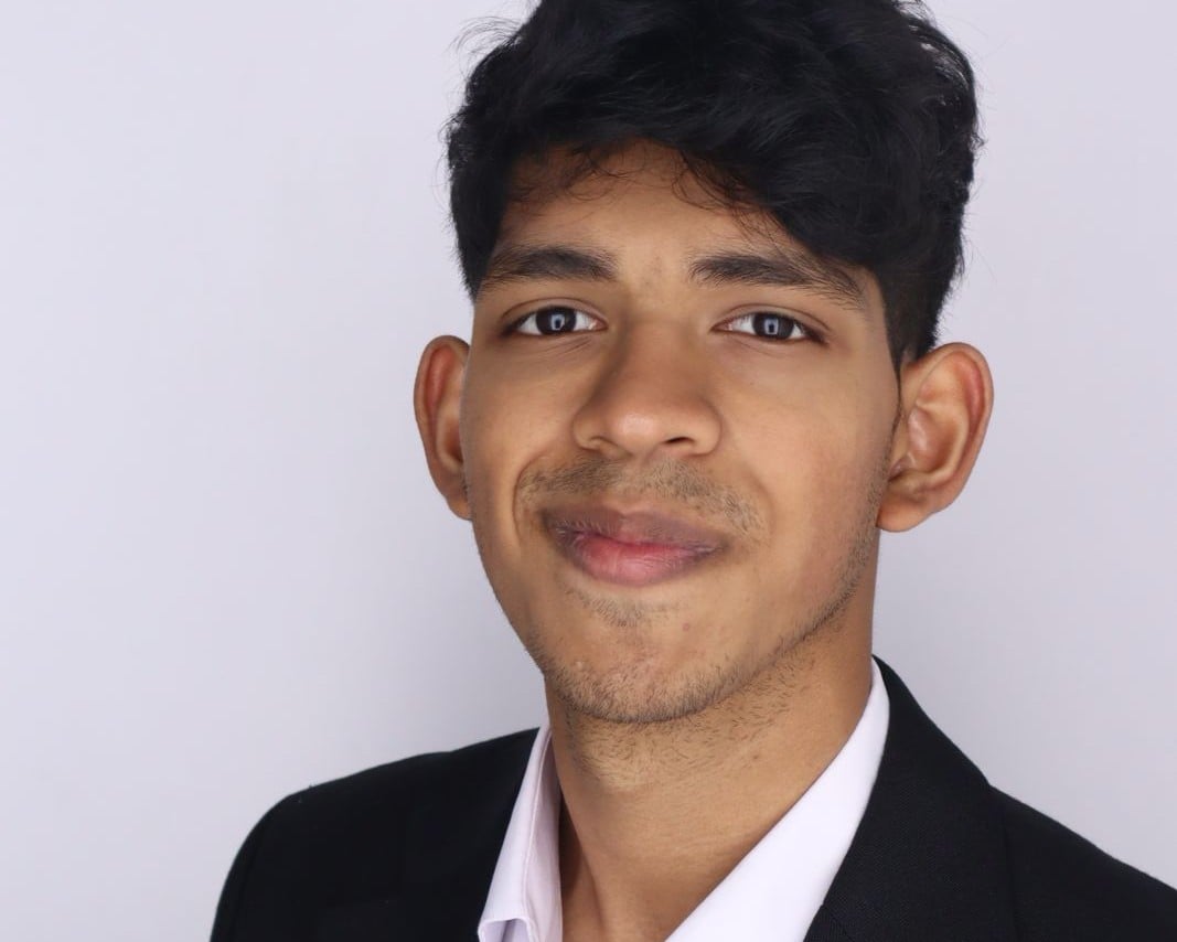 Dev Iyer with short dark hair, wearing a black blazer and a white shirt, smiles slightly while looking at the camera against a plain light background.