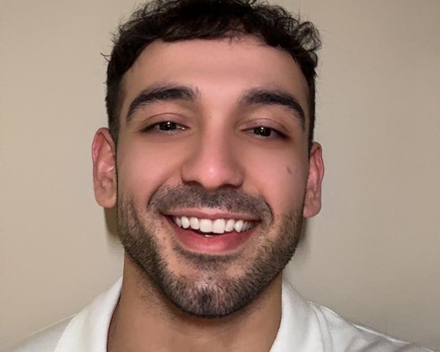 Kerem Asa with short dark hair and facial hair smiles at the camera, wearing a white collared shirt against a plain beige background.
