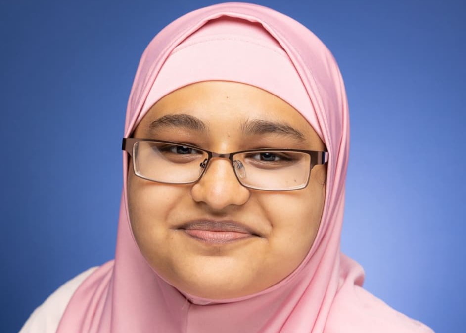 Samira Chowdhury wearing glasses and a light pink hijab smiles gently in front of a solid blue background.