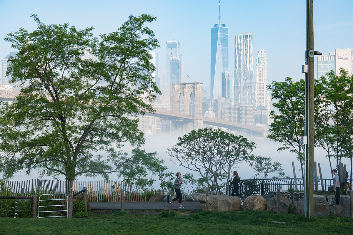 People walk along a park path with trees in the foreground. Behind them, the Brooklyn Bridge and New York City skyscrapers rise above a layer of fog on a clear morning.