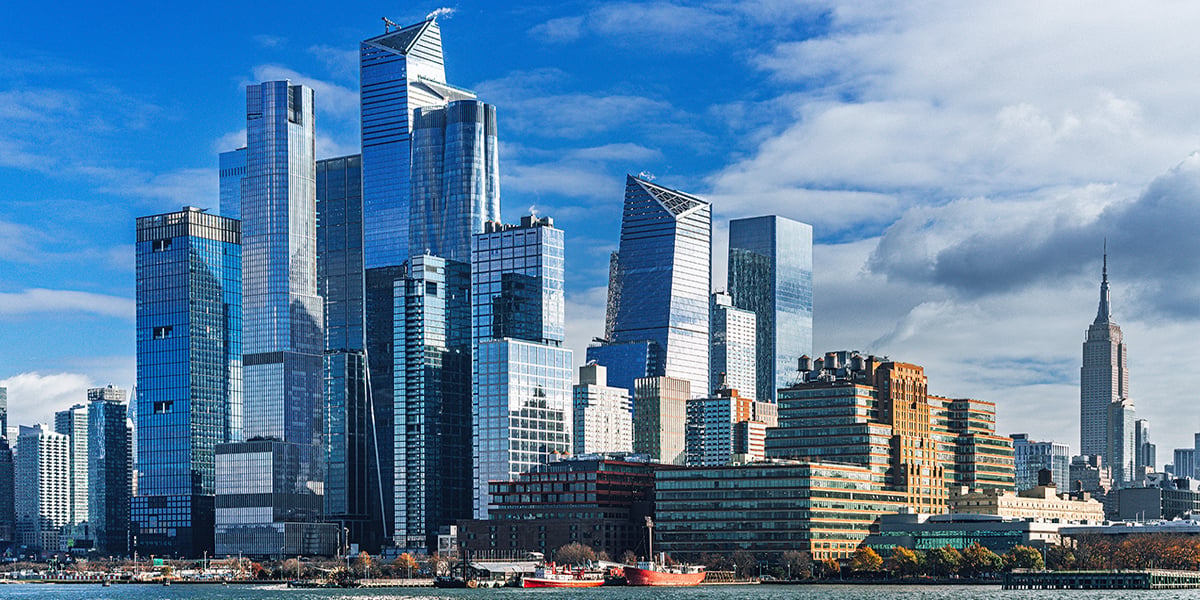 A city skyline with modern glass skyscrapers and older brick buildings, set against a partly cloudy sky. The Empire State Building is visible in the background. A river runs in the foreground.