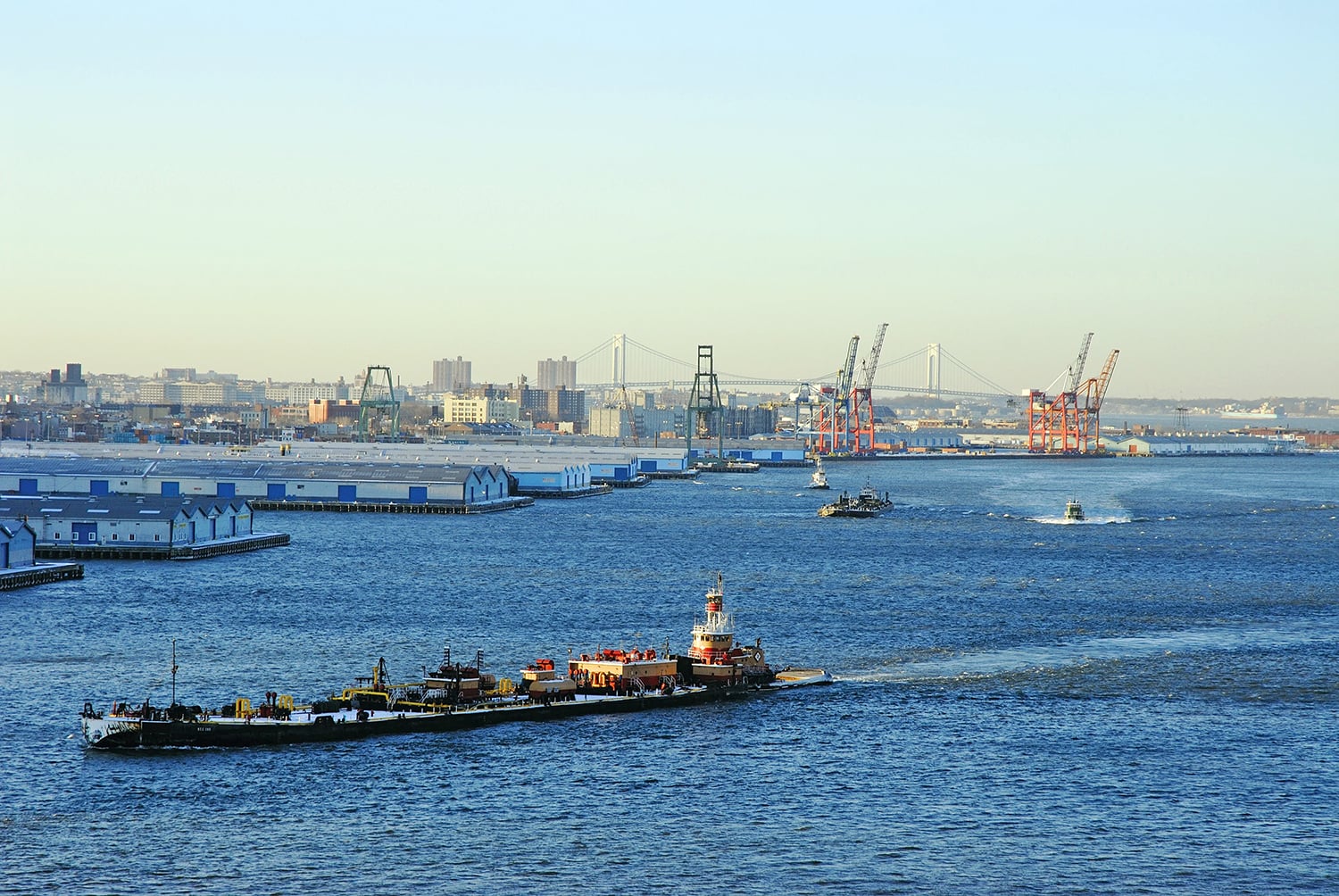 Cargo ships and tugboats move through a busy harbor with large cranes and shipping containers on the waterfront; a New York City skyline and a suspension bridge are visible in the background under a clear sky.