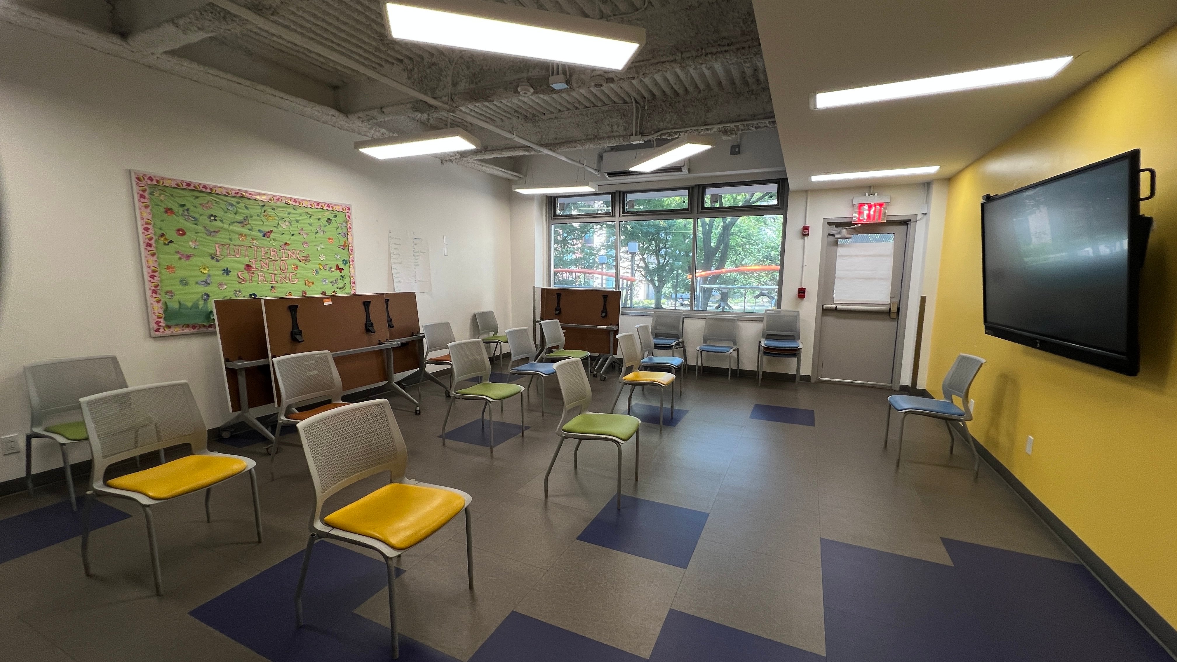 A classroom with spaced-out chairs, some with yellow seats, facing a large wall-mounted screen. There are windows and a door at the back, a bulletin board on the left, and cubby desks along one wall.