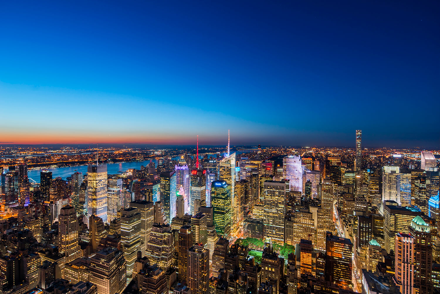 Aerial view of a brightly lit city skyline at dusk, with tall skyscrapers, illuminated buildings, and a river visible in the distance under a deep blue sky.