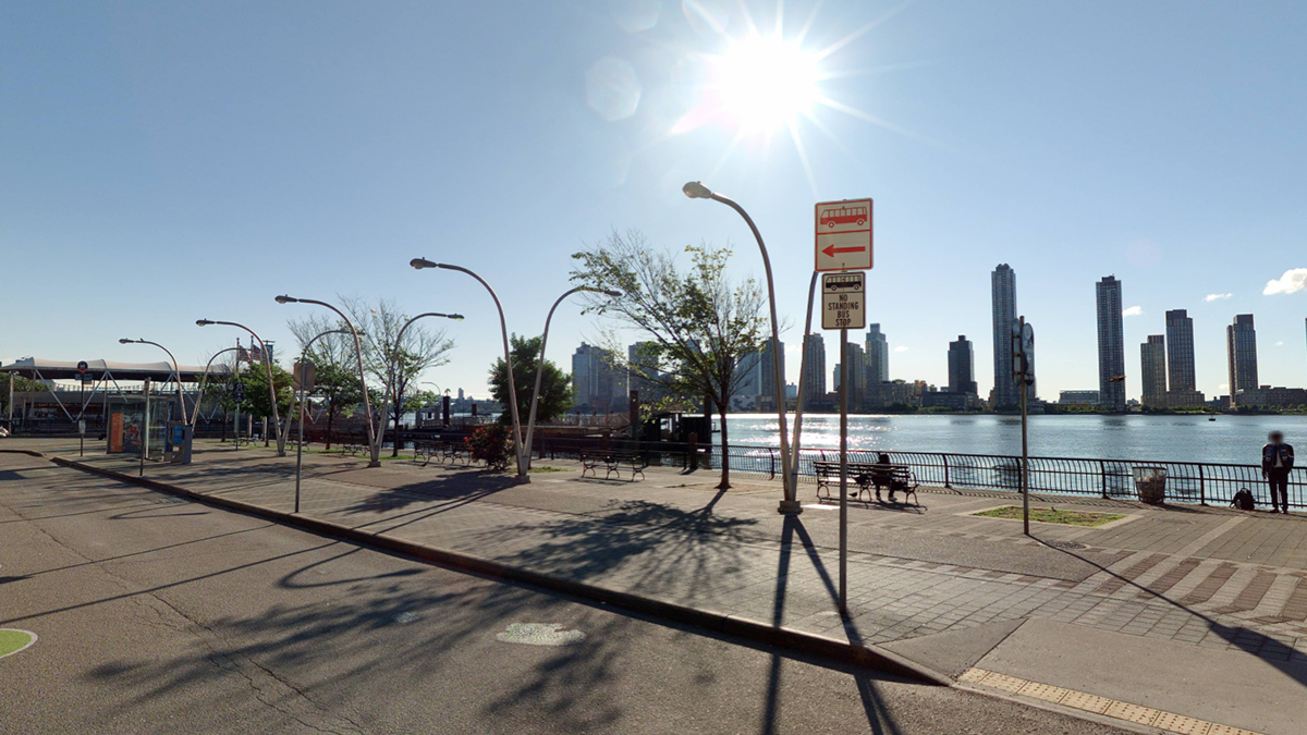 A sunny NYC scape featuring a river, tall buildings in the background, trees, street lamps, and empty benches along a riverside promenade with a few people scattered around.