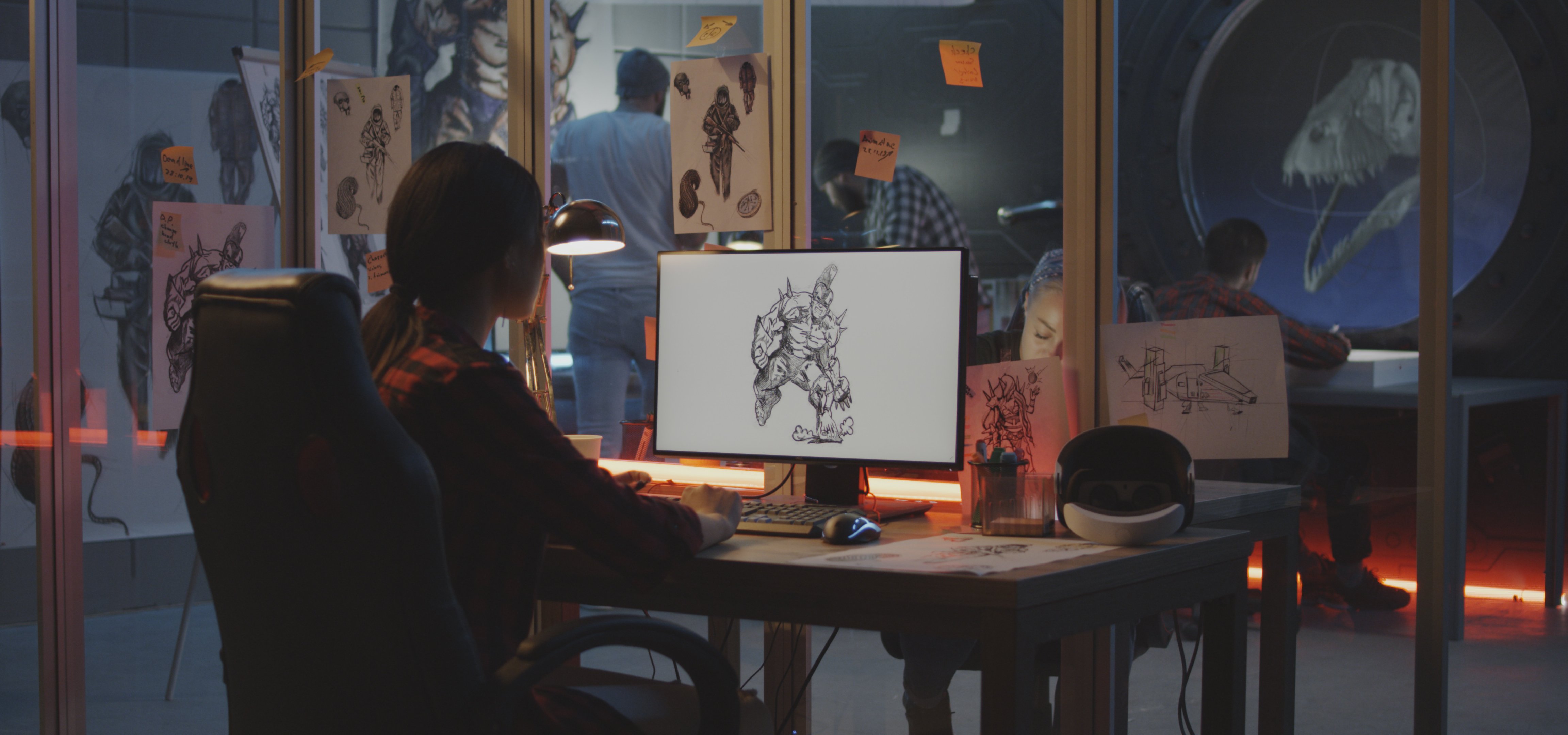 Woman sits at a desk, working on a digital drawing of a creature on a computer screen. The workspace is filled with sketches, a VR headset, and sticky notes. Other people work in the background behind glass walls.