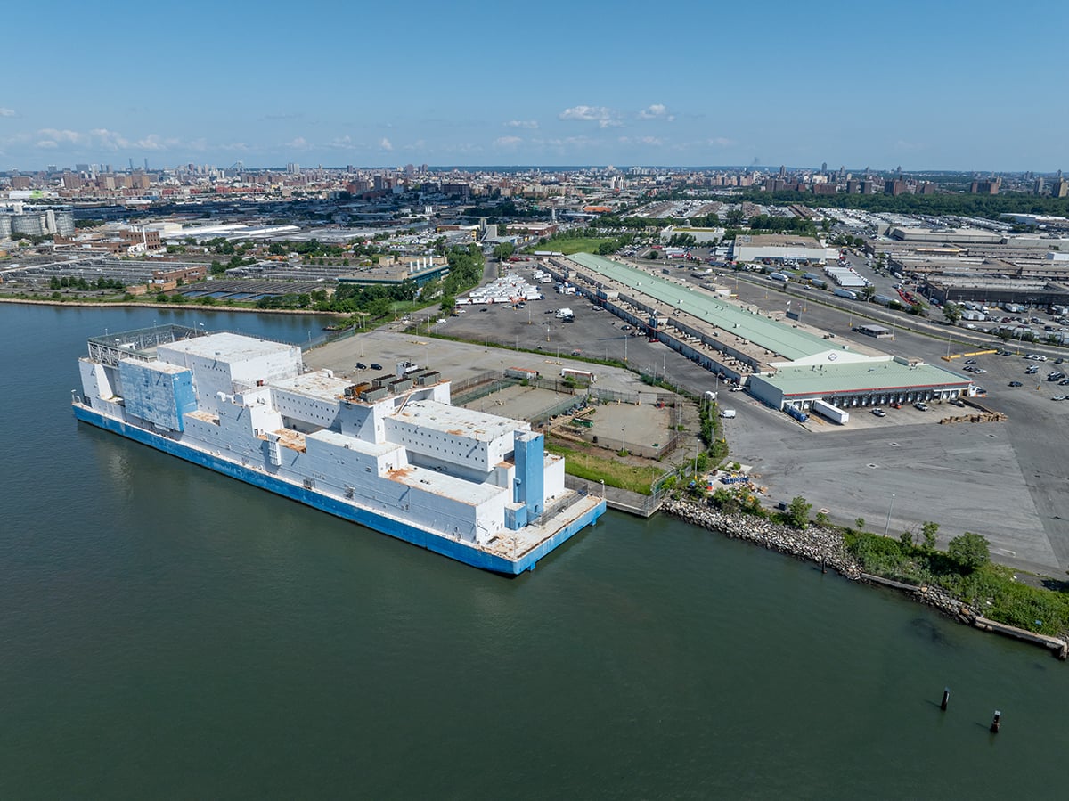 A large white and blue barge is docked at an industrial port area, with warehouses, shipping containers, and city buildings visible in the background, under a clear blue sky.