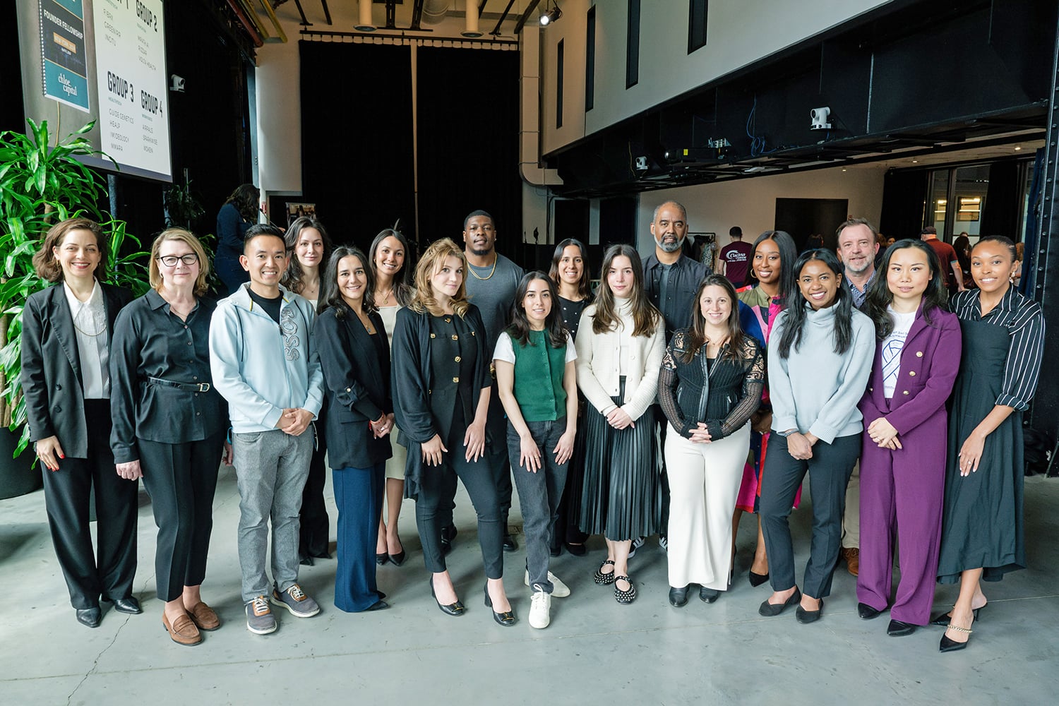 A diverse group of 18 people, both men and women, stand together indoors, posing and smiling for a group photo. They are dressed in business and business-casual attire.