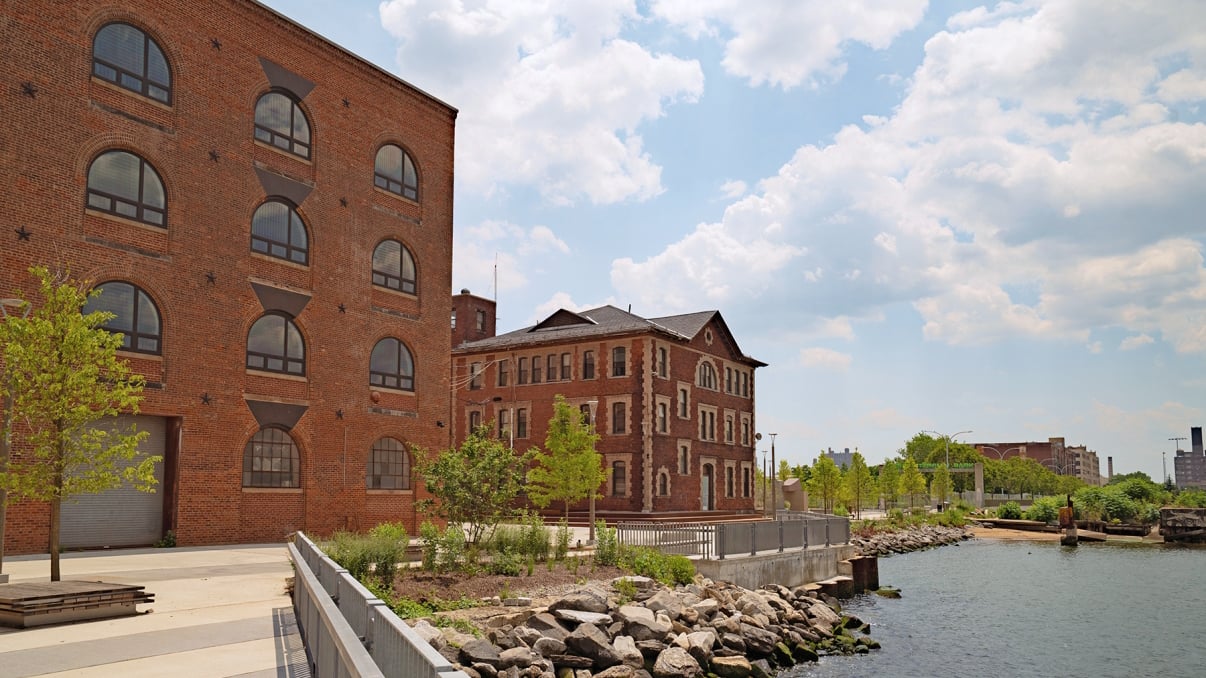 Red brick industrial buildings sit beside a river, surrounded by trees and rocks along the water’s edge. The sky is partly cloudy, and the area appears calm and well-maintained.