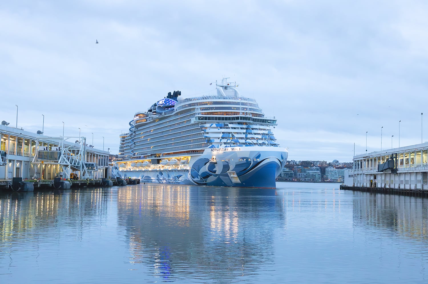 A large cruise ship, the Norwegian Prima, between two piers at the Manhattan Cruise Terminal on a calm, cloudy day. The ship features blue and white designs on its bow and is illuminated with lights. Buildings are visible in the background.