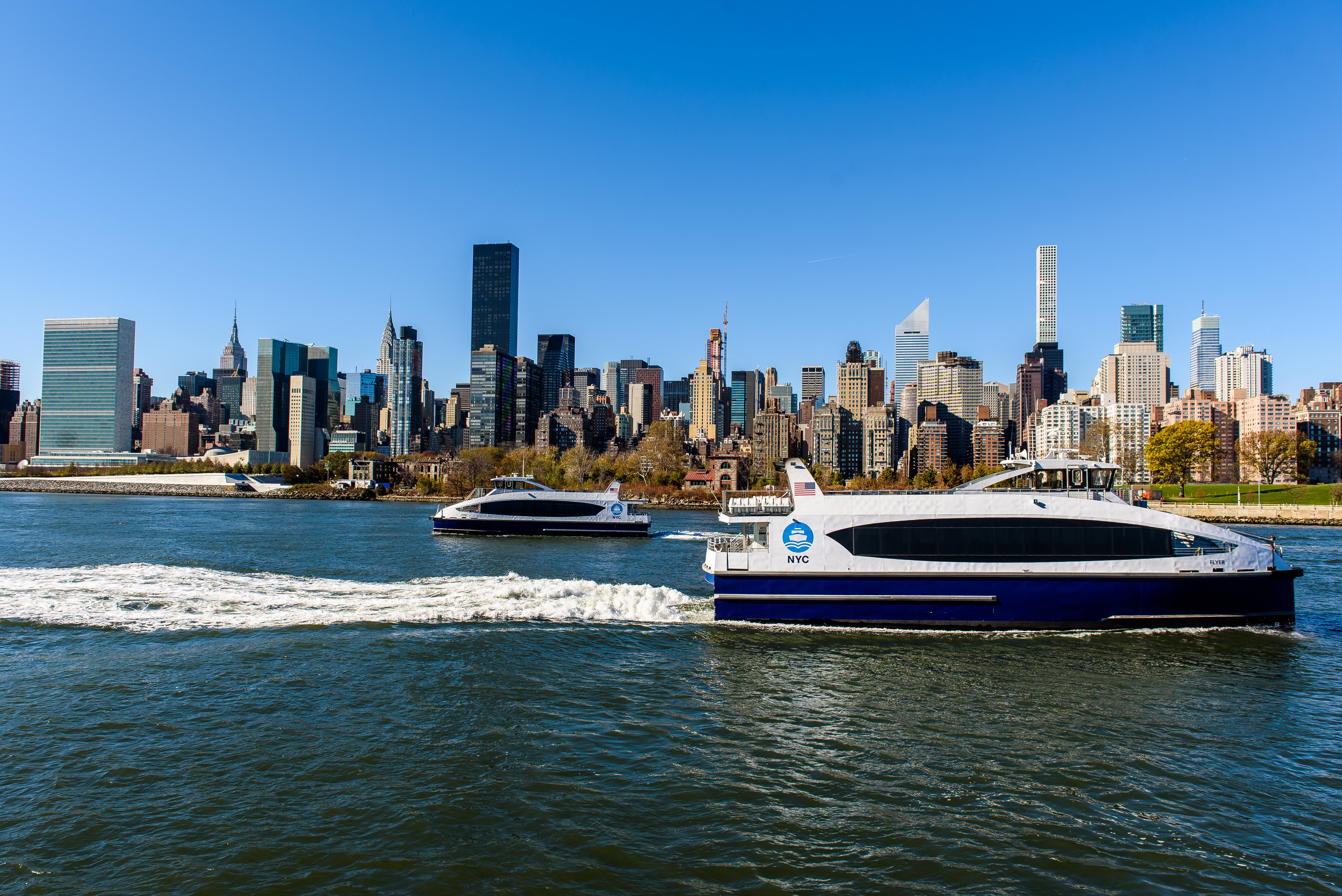 Two NYC ferries travel on the East River with the Manhattan skyline, including tall skyscrapers and clear blue sky, in the background.