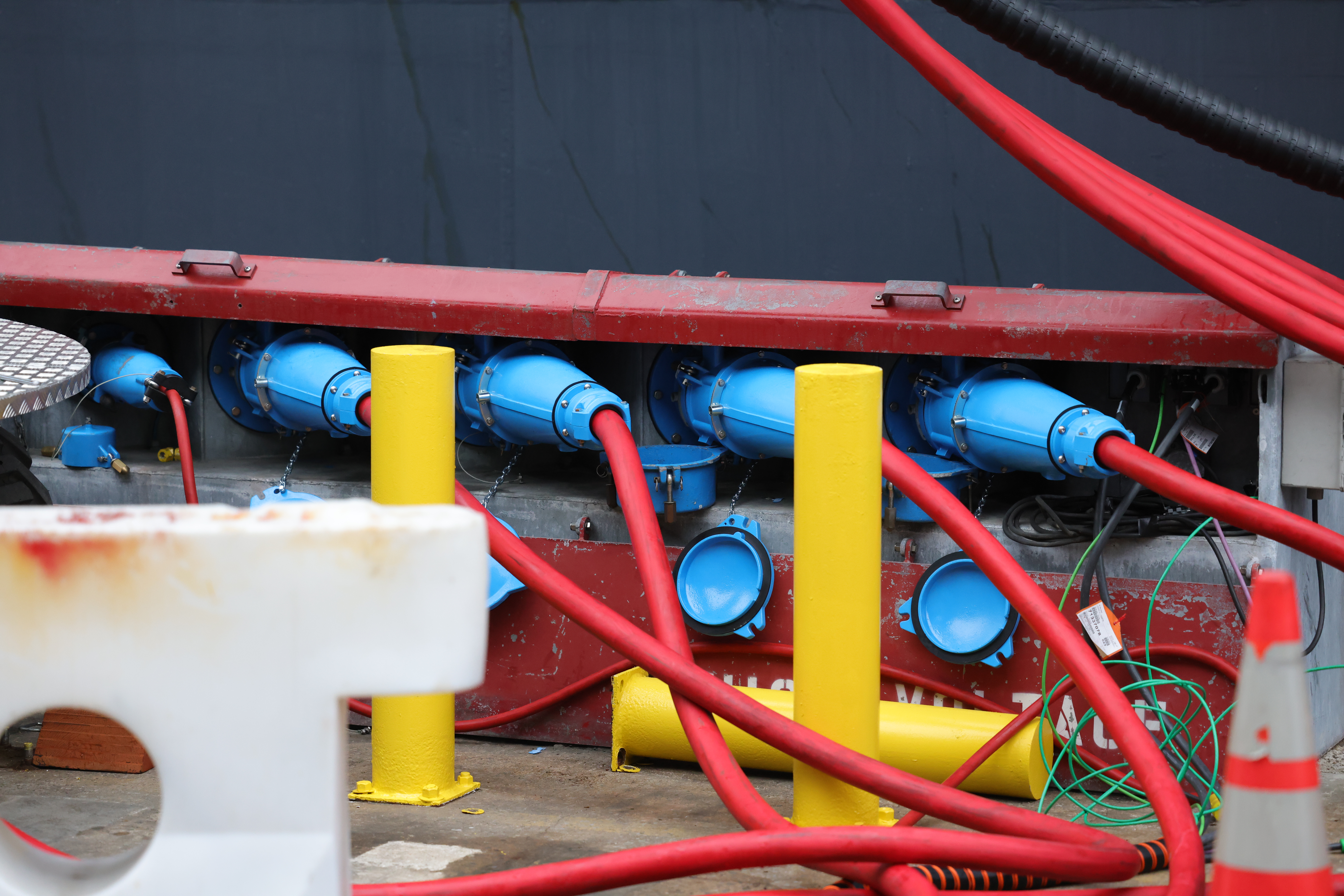 Multiple thick red cables are connected to blue industrial connectors mounted on a metal panel. Yellow protective posts are in front, and green wires and an orange safety cone are nearby. The background is a dark wall.