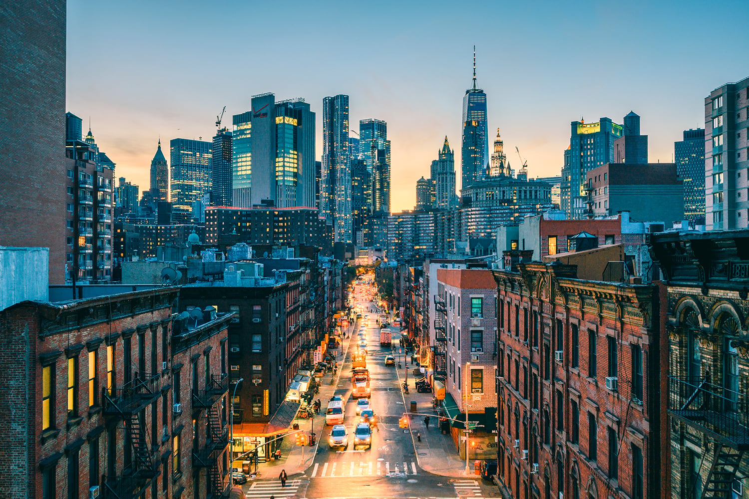 A New York City street lined with brick buildings stretches toward a skyline of tall, modern skyscrapers at sunset. Cars drive along the lit street, while the sky glows with warm evening light.