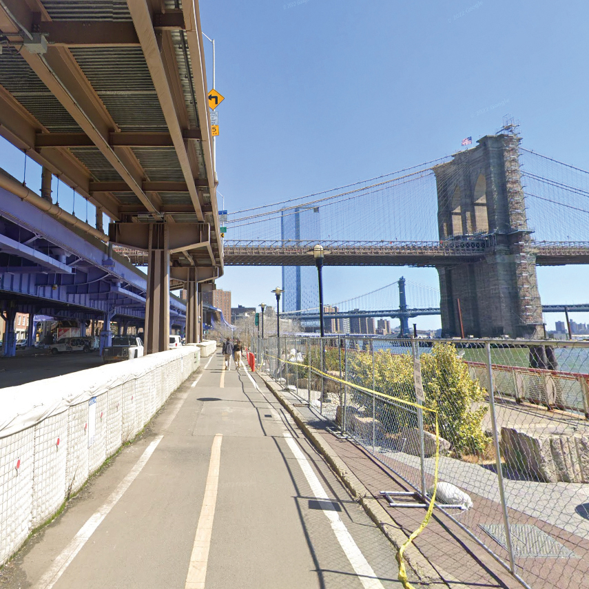 A pedestrian and bike path runs alongside a fenced-off area and elevated road, with the Brooklyn Bridge and Manhattan Bridge visible in the background under a clear blue sky.