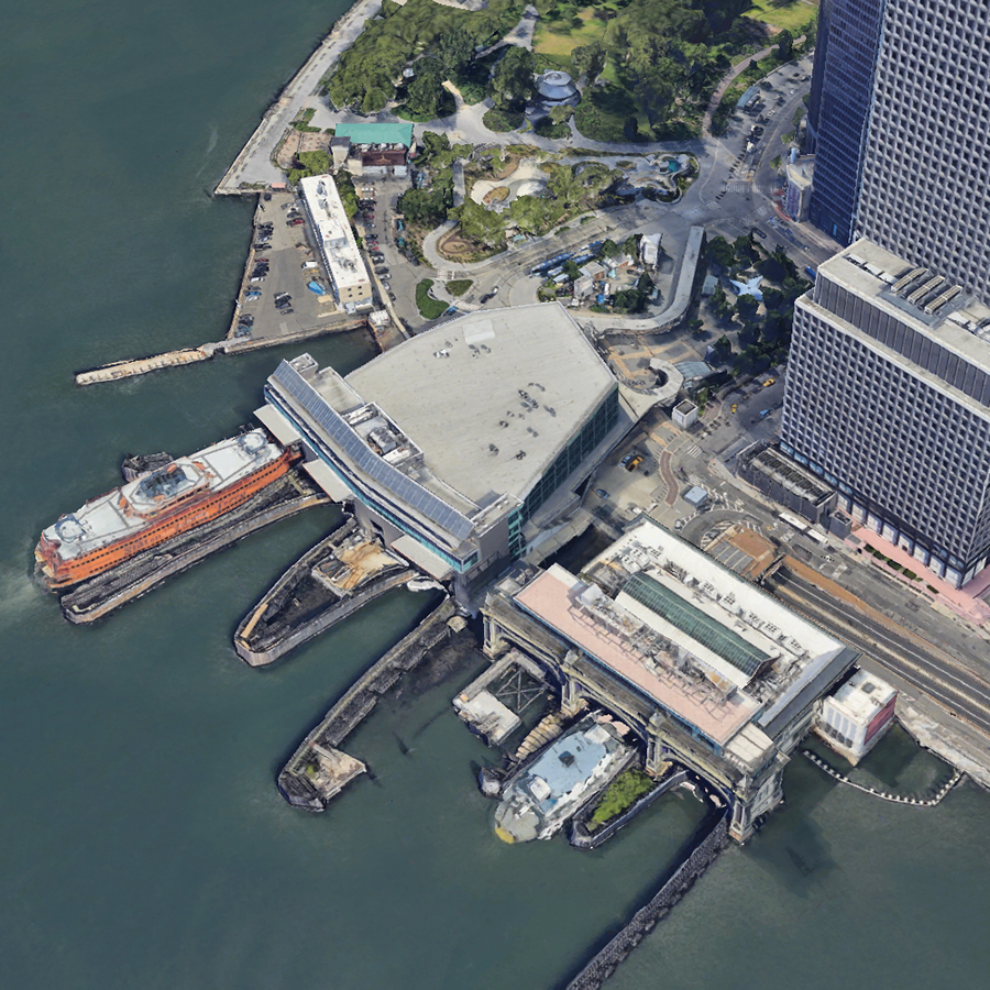 Aerial view of Whitehall Ferry Terminal, including the Staten Island Ferry, the Battery Maritime Building, and Governors Island Ferry. Ferries docked by the waterfront, next to tall office buildings and a nearby park with trees and pathways.