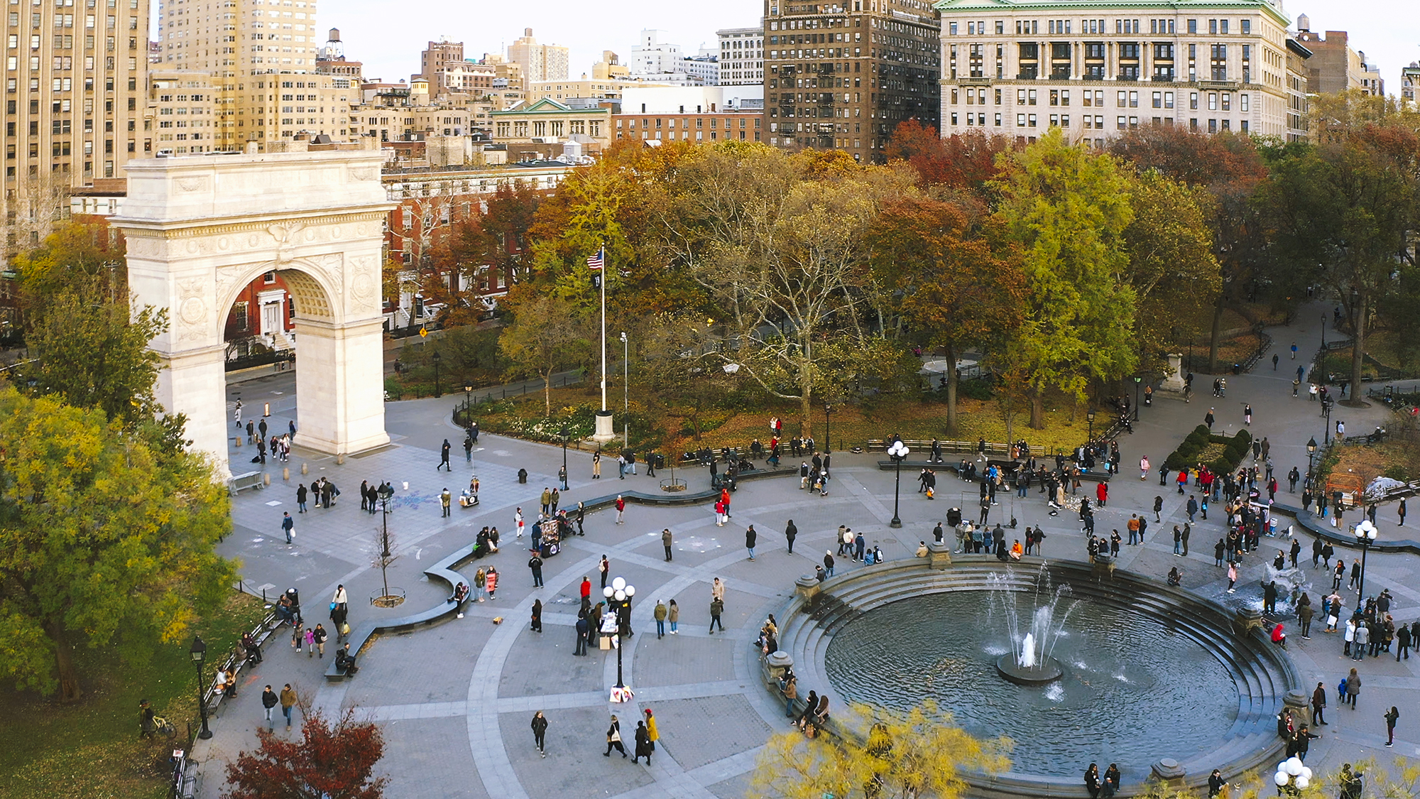 Aerial view of Washington Square Park in New York City with people walking near the arch and around the central fountain, surrounded by autumn trees and city buildings.