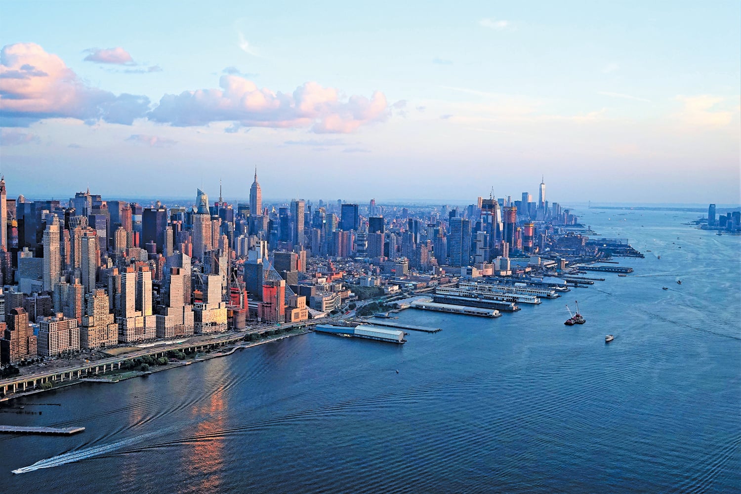 Aerial view of Manhattan, New York City at sunset, showing skyscrapers, waterfront docks, and boats moving on the Hudson River under a partly cloudy sky.