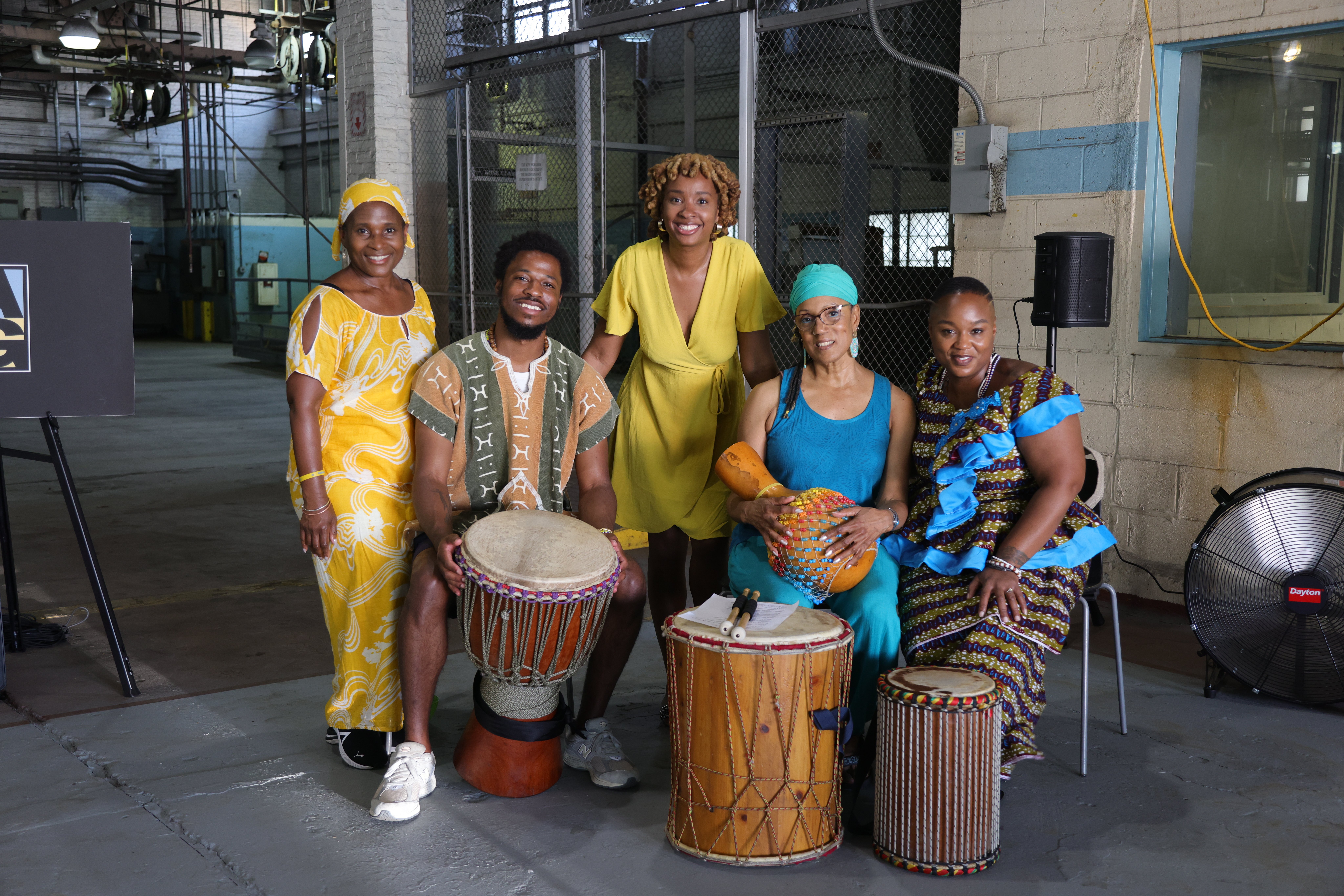 Five people in colorful, traditional African attire pose indoors with various drums and percussion instruments, smiling at the camera. The setting is an industrial-style room with exposed pipes and a large fan.