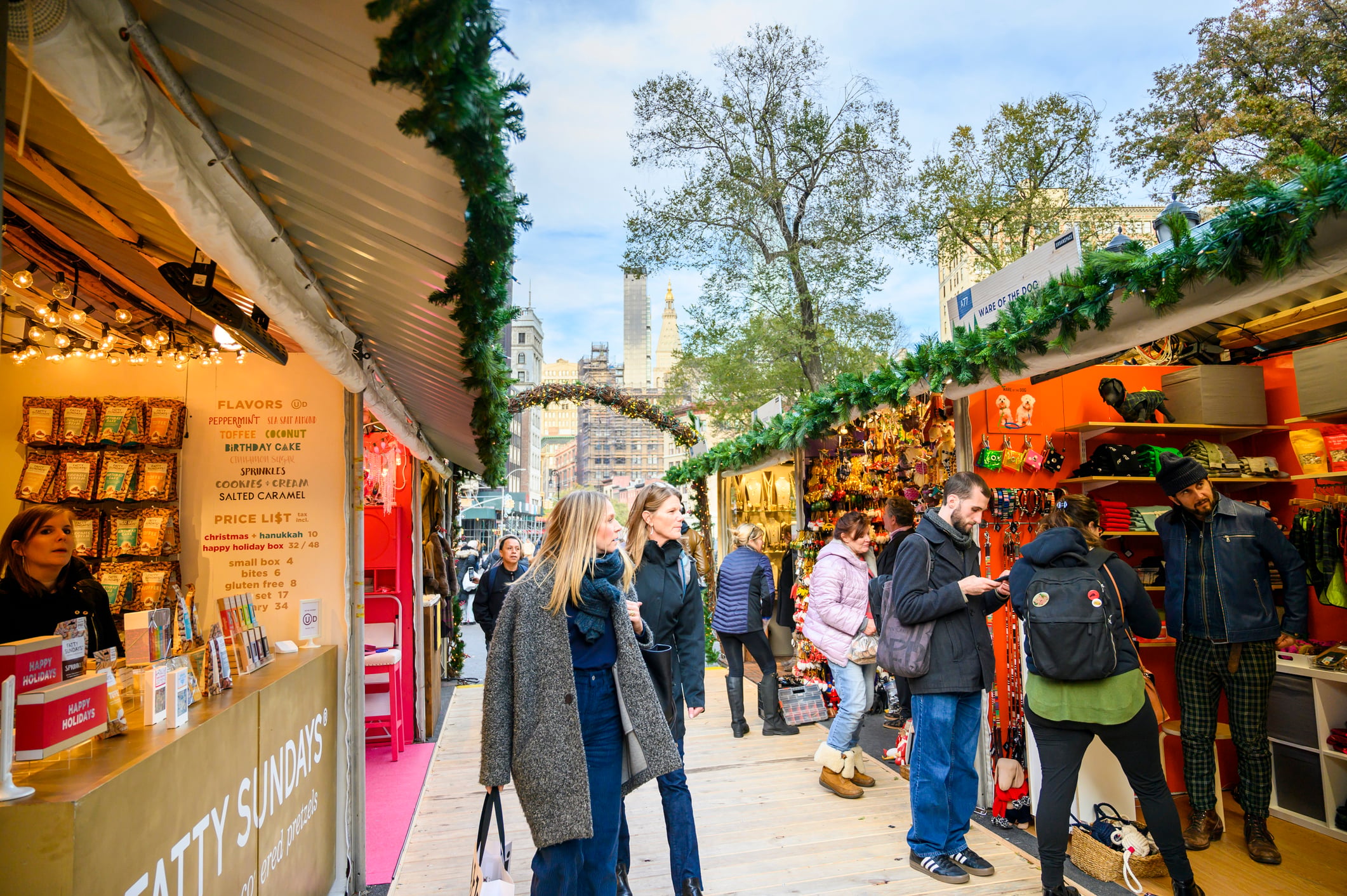People browse festive Union Square Holiday Market stands decorated with greenery and lights. Vendors display snacks, gifts, and holiday items. Trees and tall buildings are visible in the background under a bright sky.