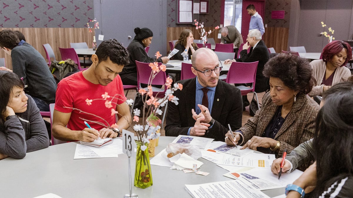 A group of diverse people at the Non-Profit Coffee hour sit around a round table in a community setting, engaged in discussion. Papers, pens, and a vase with flowers are on the table. Other people are visible in the background, seated at additional tables. Photo by Kreg Holt/NYCEDC.