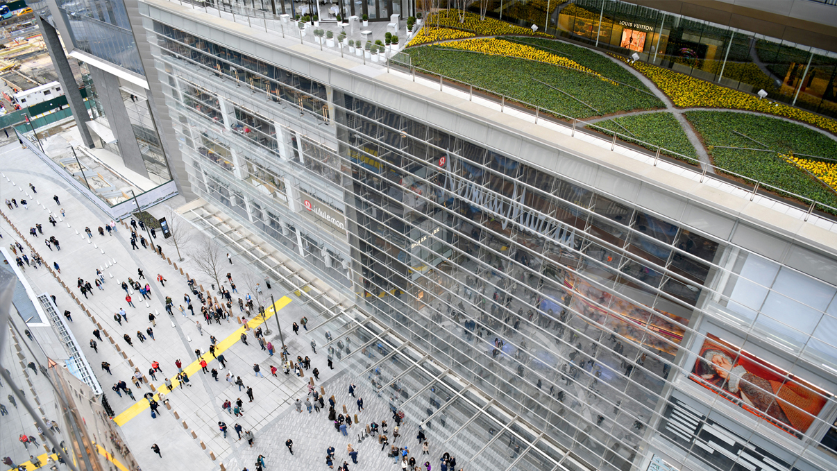 Aerial view of Hudson Yards Shopping, a modern and large building with a glass facade and a green rooftop garden. People are gathered on the spacious, light-colored pavement below. The buildings architectural details and outdoor space are prominently visible. Photo by Clint Spaulding/Getty Images.