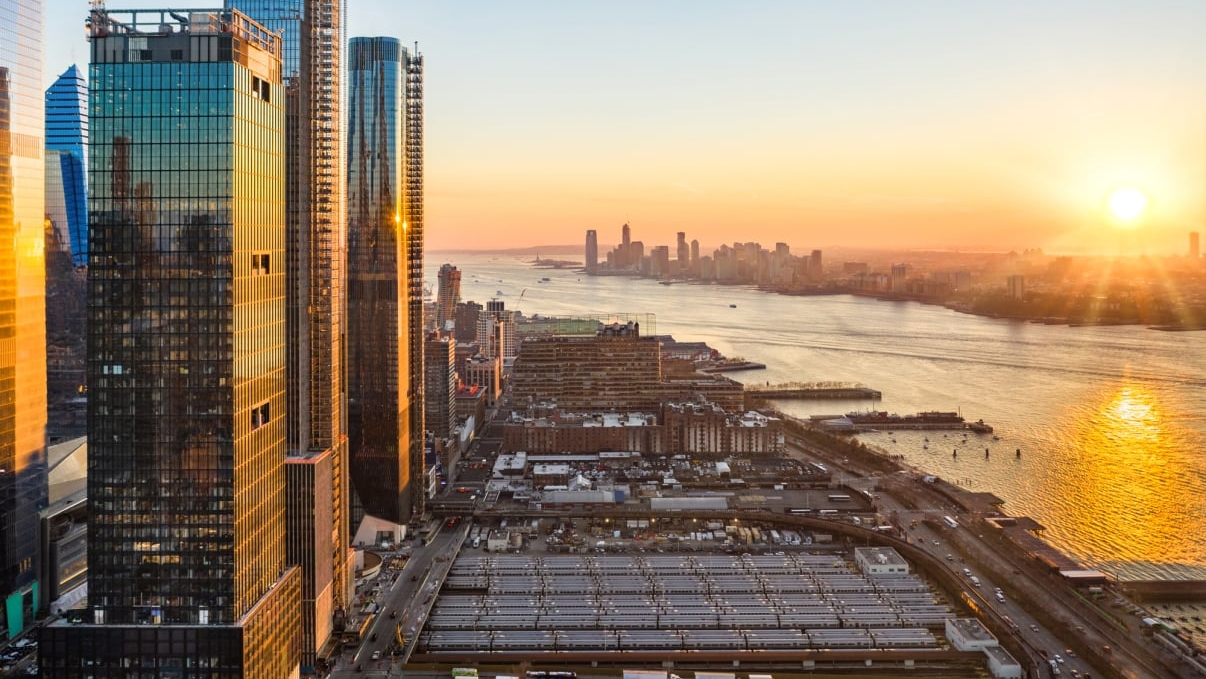View from Hudson Yards of New York City skyline at sunset, featuring tall skyscrapers reflecting the suns orange glow. A river runs through the city, with boats visible on the water. The sky is clear, with the sun setting near the horizon.