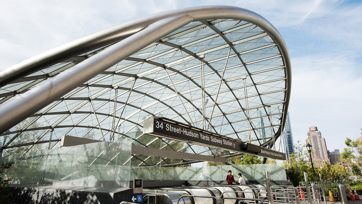 34th St-Hudson Yards Subway Station entrance with a large, curved glass and steel canopy. Escalators lead down, with buildings visible in the background. Skies are partly cloudy. Photo by Julienne Schaer.