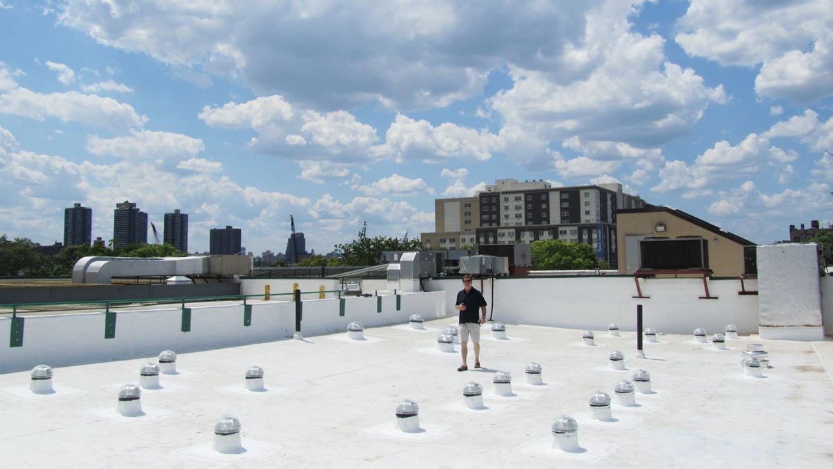 A person stands on a white rooftop scattered with small vents. The background shows NYC skyline with high-rise buildings and fluffy clouds against a bright blue sky.