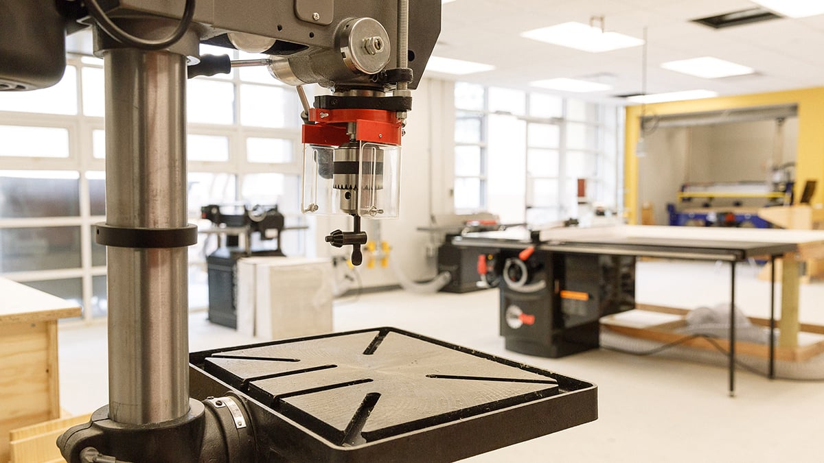 An industrial workshop with a large drill press in the foreground. The room is spacious and well-lit with large windows. In the background, there is a workbench and various tools and equipment scattered around. The walls are yellow and white. Photo by Kreg Holt/NYCEDC.