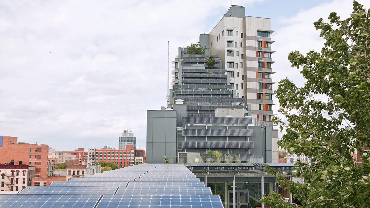 A modern building with multiple balconies covered in greenery, set against a cityscape. In the foreground, solar panels are installed on a roof, reflecting the cloudy sky. Trees are visible on the right, adding to the urban green space.