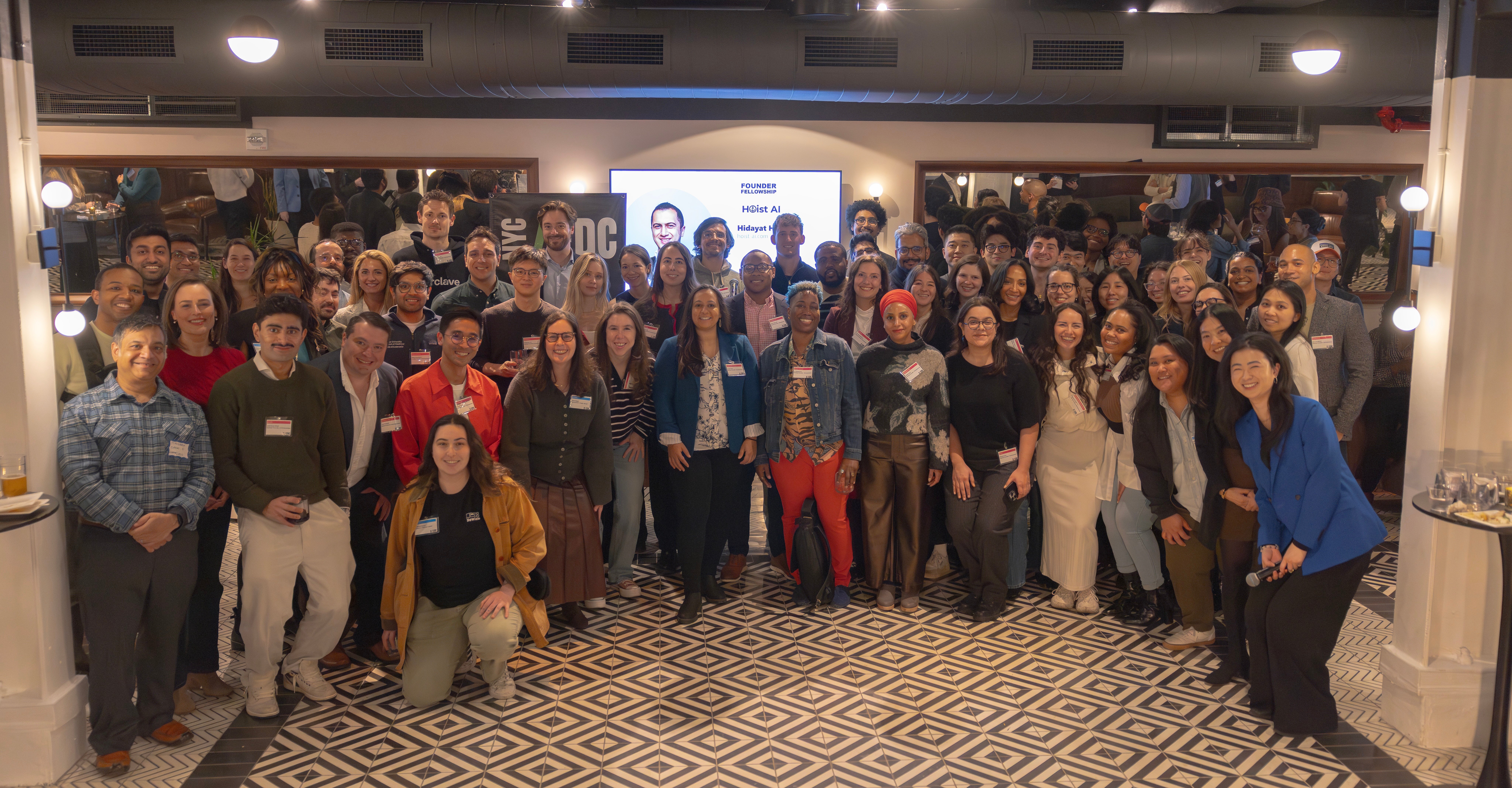 2026 Founder Fellowship cohort pose and smile for a group photo in a well-lit indoor space with patterned floors, mirrors, and a screen in the background. Most of them are wearing name tags.