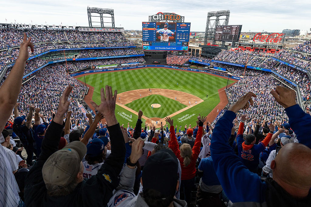 Fans cheer in a packed stadium at Citi Field during a baseball game between the New York Mets and Toronto Blue Jays, with the field, scoreboard, and NYC skyline visible in the background. Players are on the field and the atmosphere is lively and energetic. Photo by Al Bello/Getty Images.