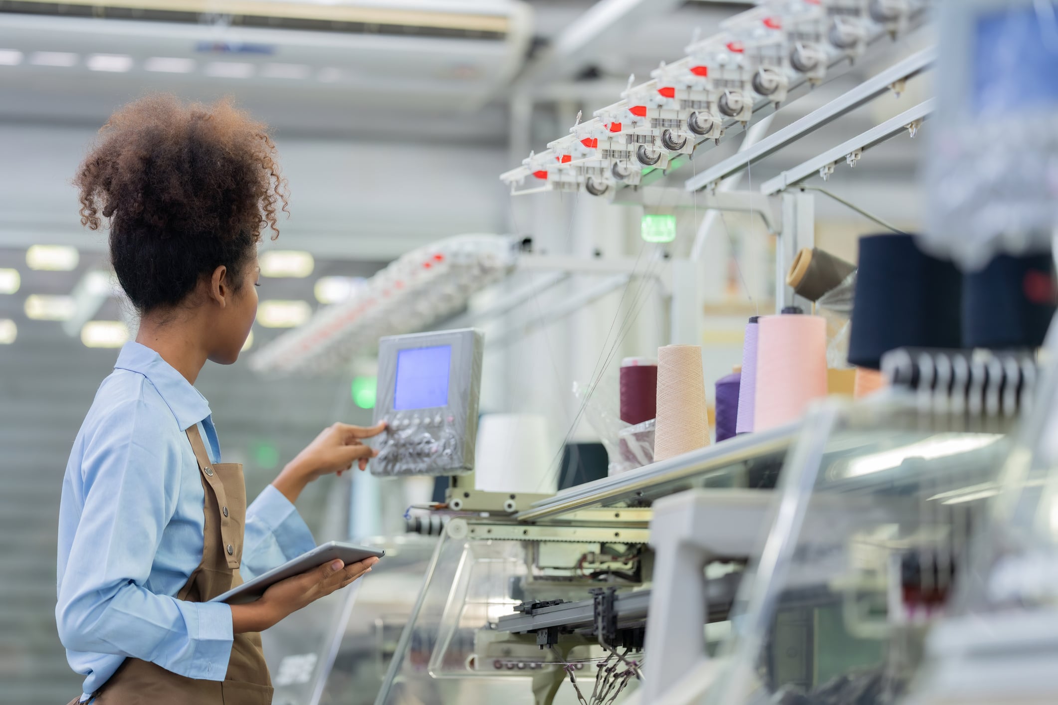 A woman wearing a blue shirt and apron operates a control panel in a modern textile factory, holding a digital tablet beside spools of colorful thread and automated machinery.