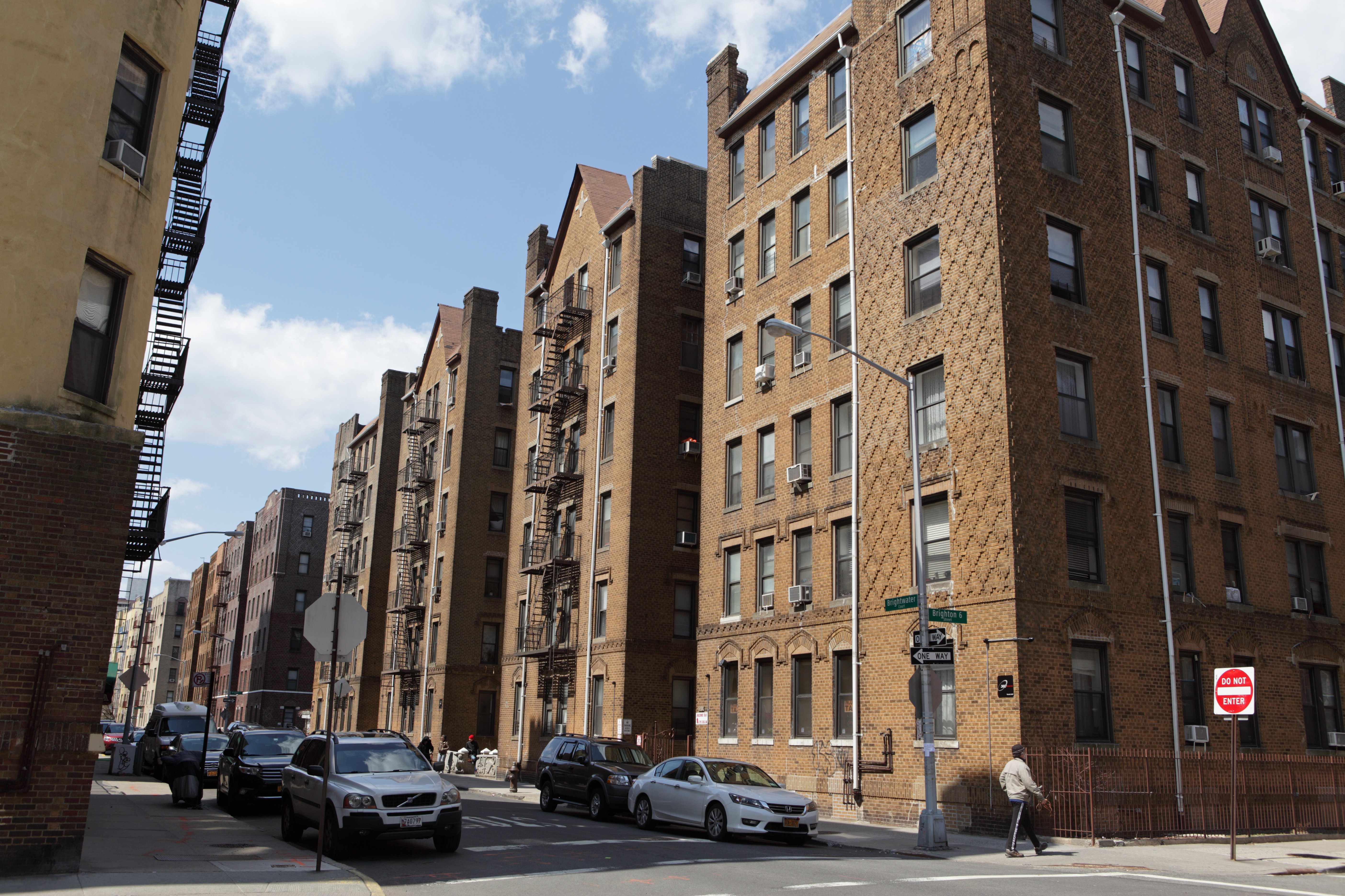A street corner in Brighton Beach, Brooklyn with parked cars and tall brown brick apartment buildings. A person with a cane is walking on the sidewalk under a partly cloudy sky.
