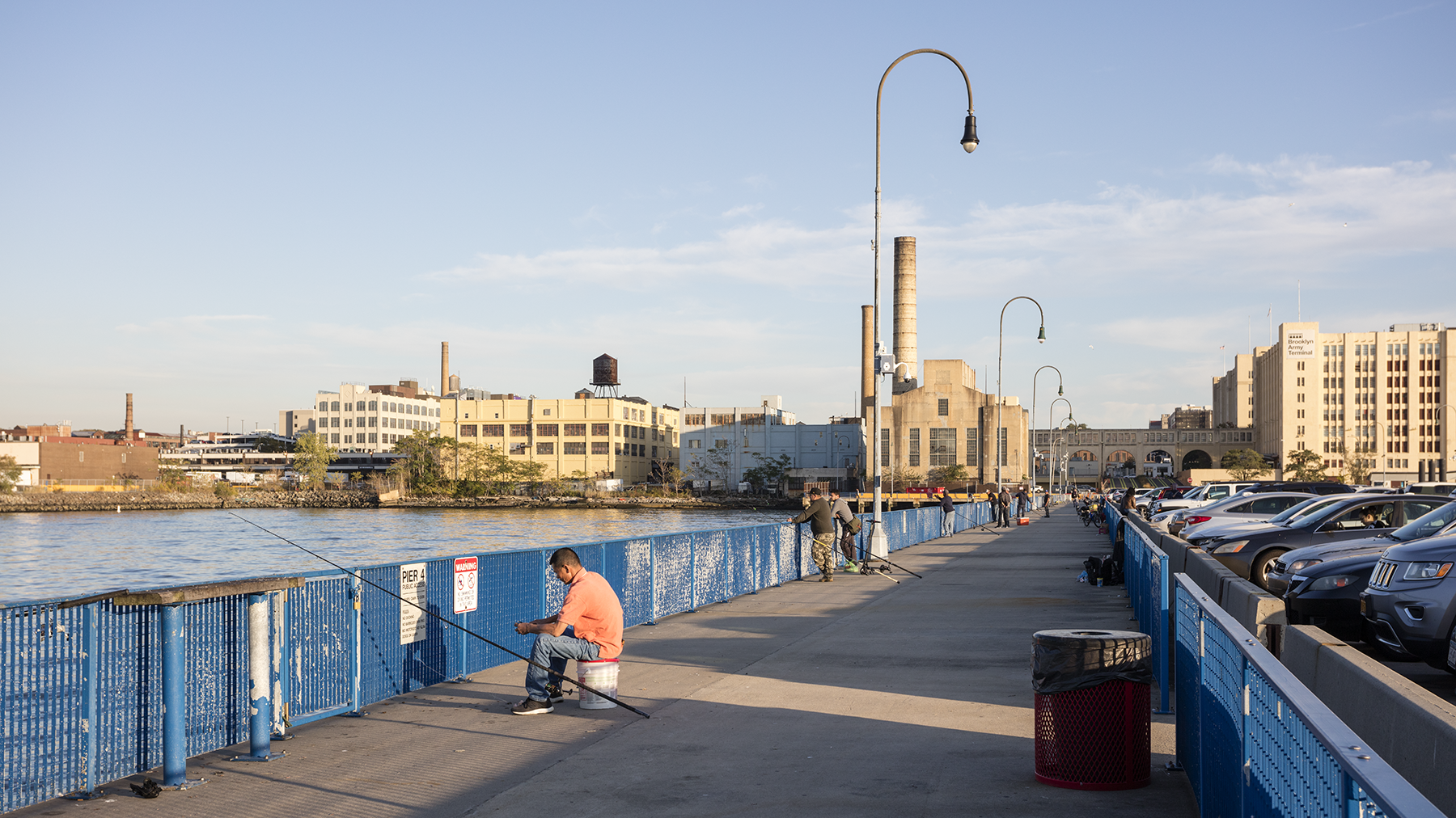 A person sits on a stool fishing on a blue metal pier under a clear sky. Nearby, people walk along the pier lined with vintage-style street lamps. Industrial buildings and parked cars can be seen in the background.
