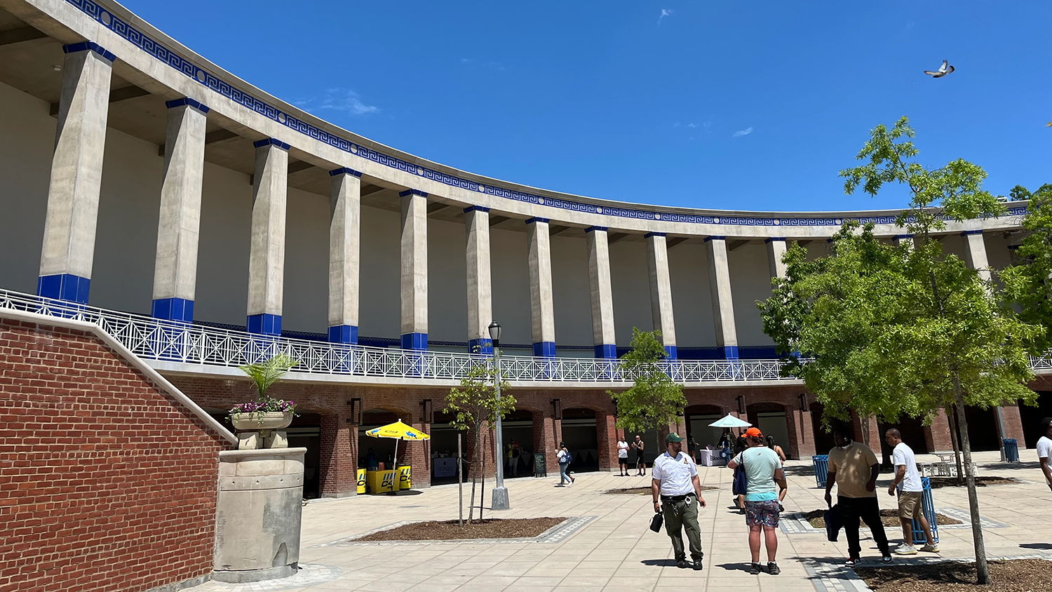 People walk in a sunny open plaza with large trees inside a curved building featuring tall columns and blue accents. A few yellow umbrellas provide shade, and a bird is flying in the clear blue sky.
