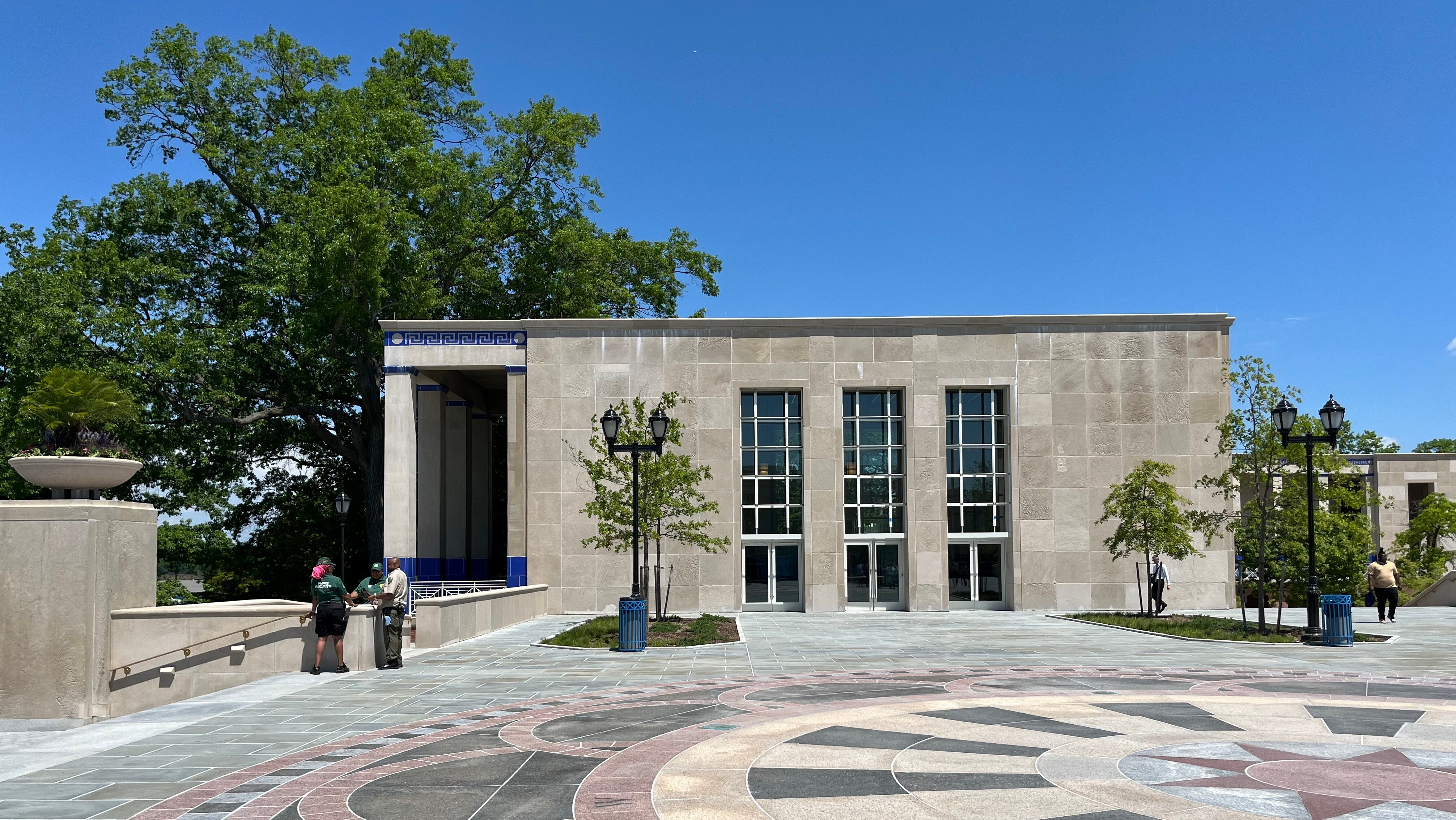 A modern stone building with large vertical windows stands behind a patterned circular plaza. Several people are gathered near the left railing, and trees frame the scene under a clear blue sky.