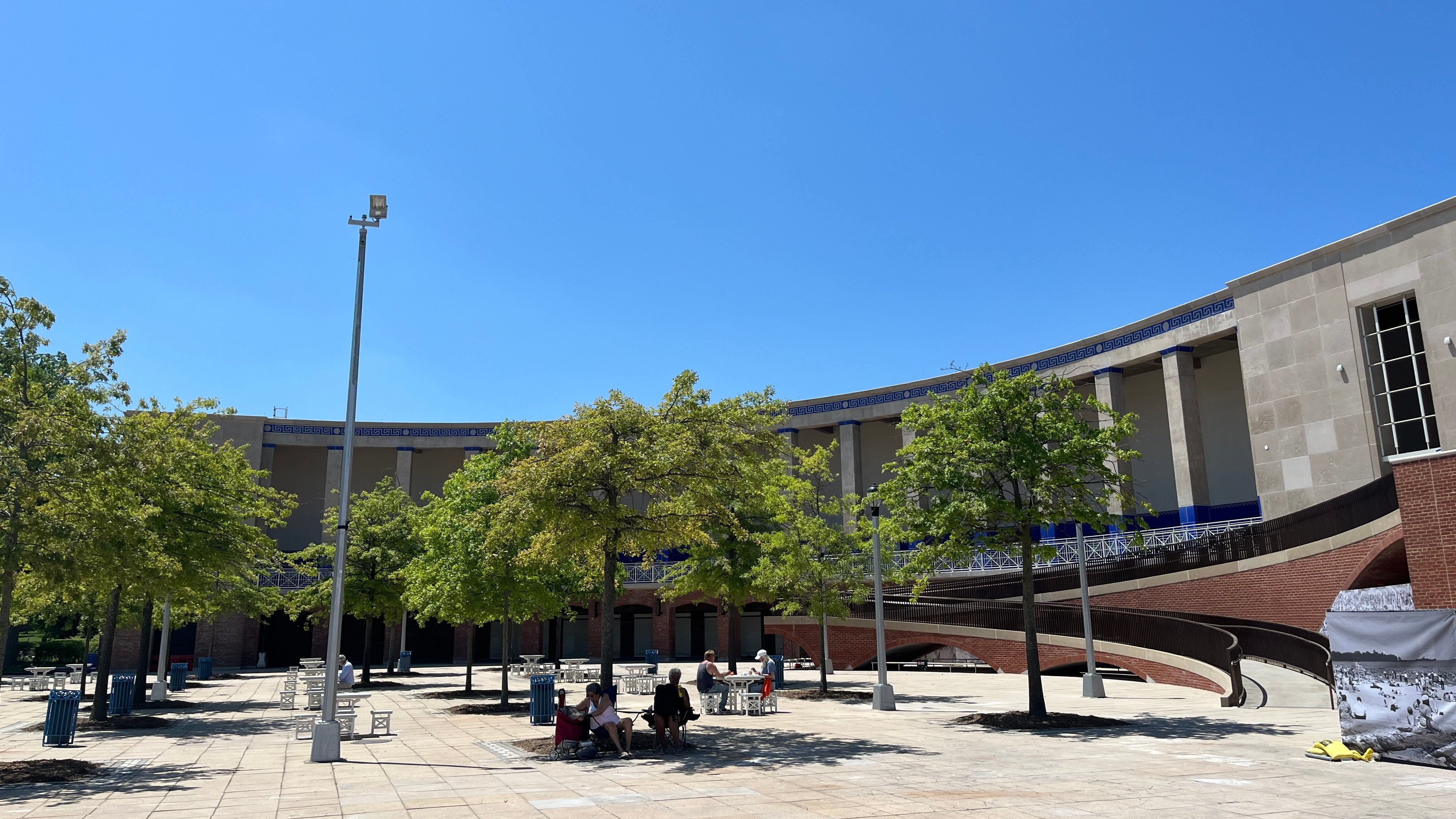 A sunny outdoor plaza with green trees, benches, and people sitting in the shade. In the background, there is a curved, columned building with stone walls and a blue sky overhead.