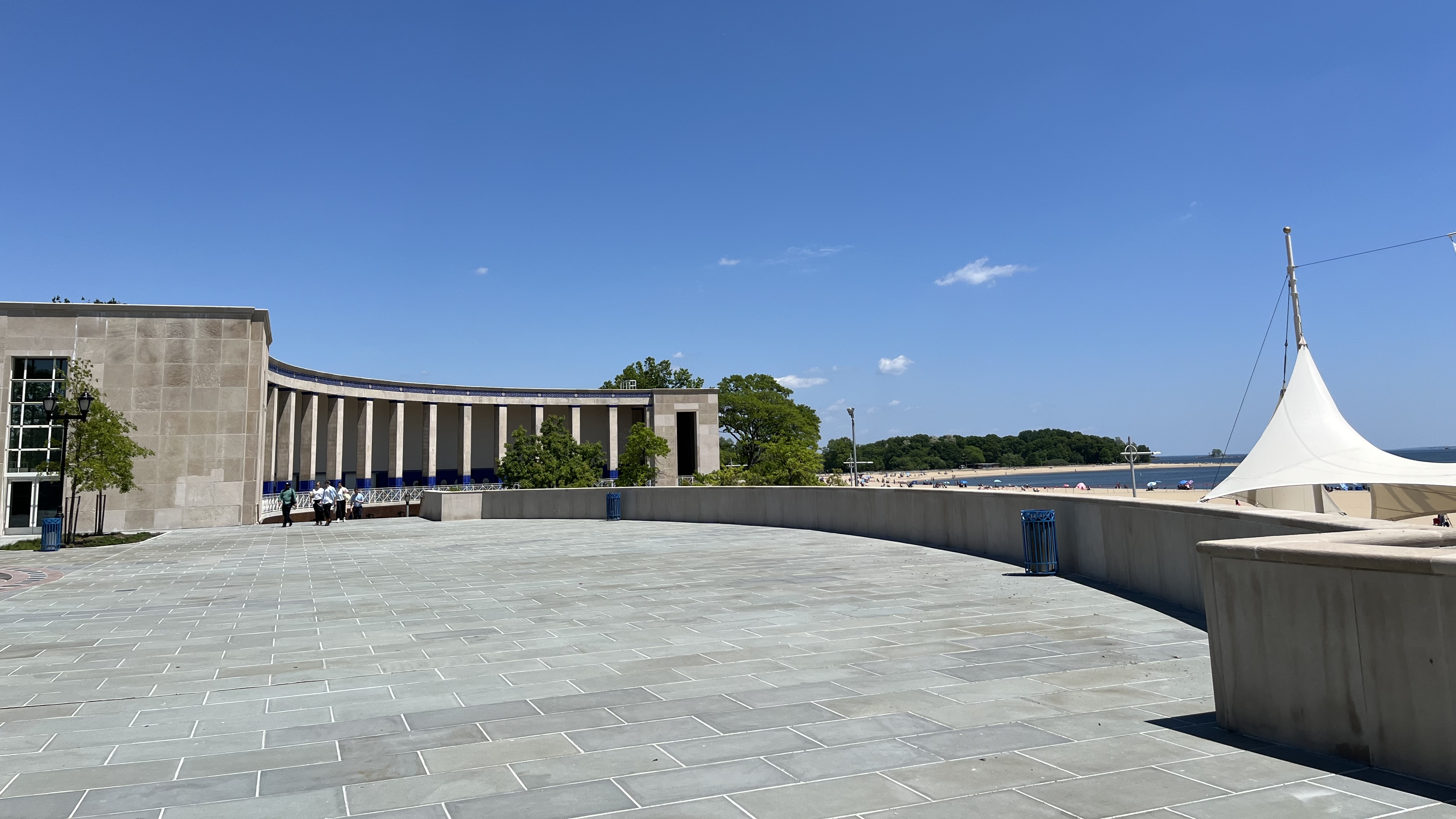 A wide stone plaza with a curved building on the left, a white canopy on the right, a few people walking, green trees, a sandy beach, and blue sky in the background.