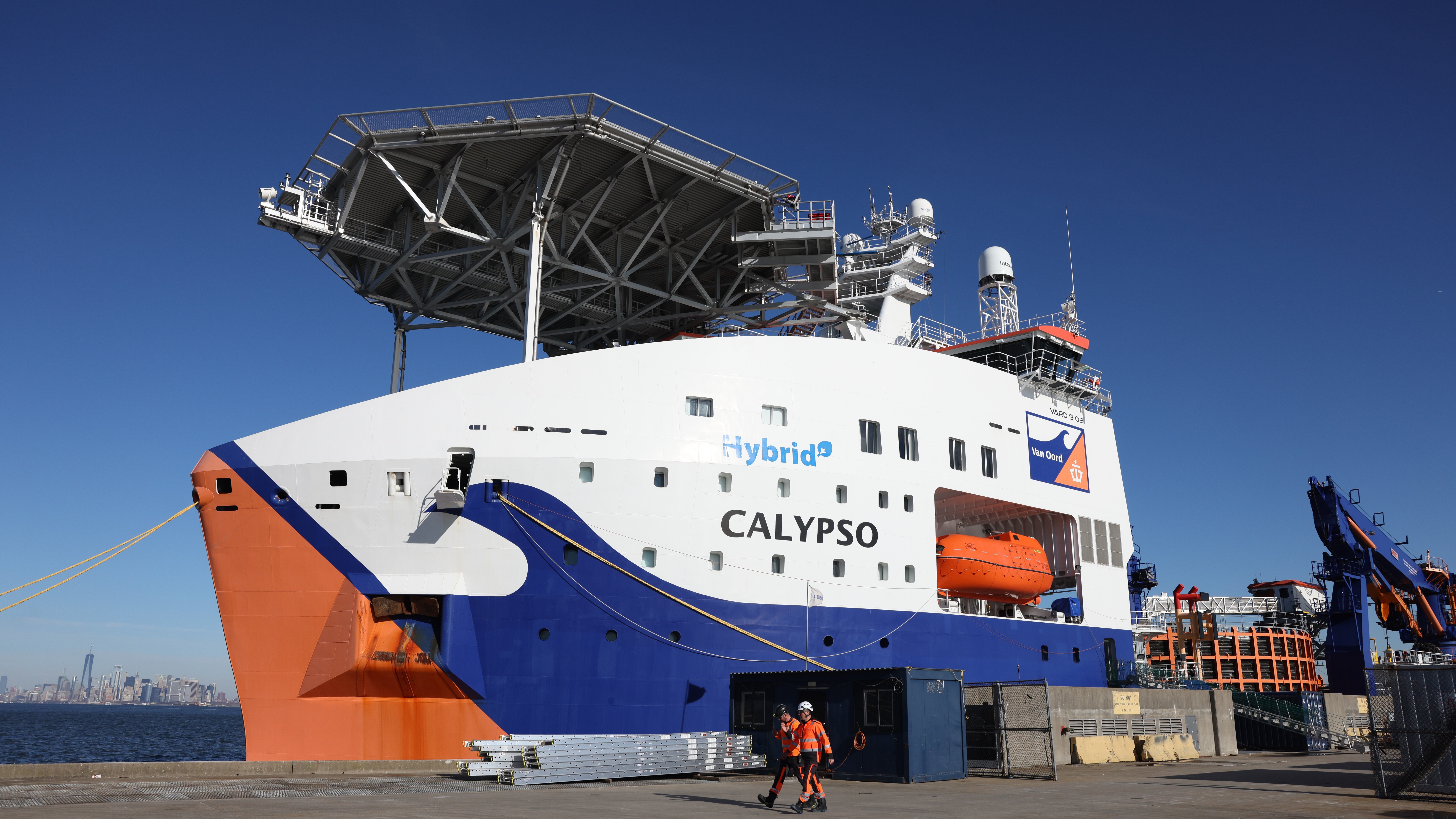 A large hybrid ship named CALYPSO docked at an NYC public pier under a clear blue sky, with two workers in orange safety gear walking nearby. The New York City skyline is visible in the distant background.