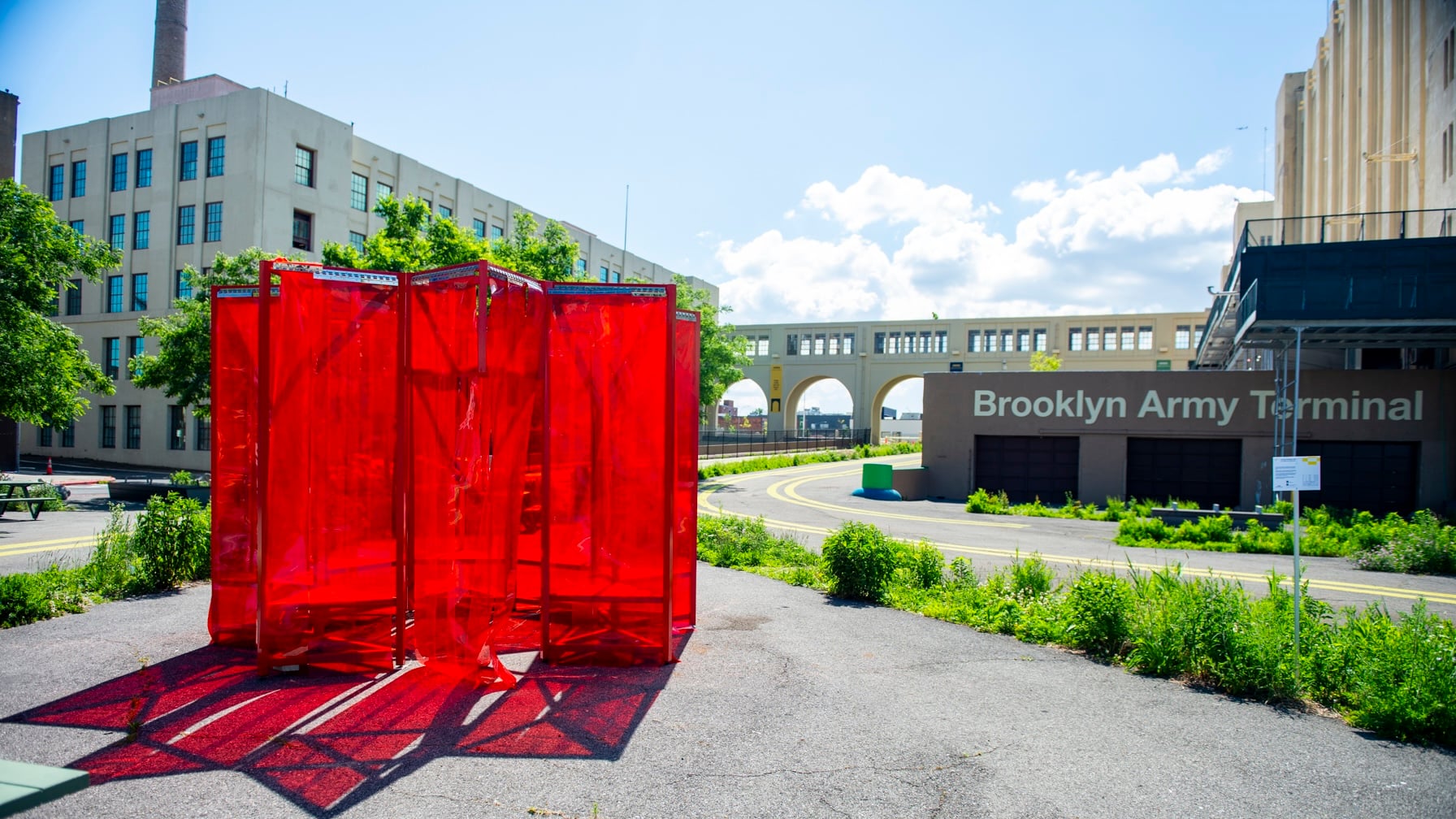 A large outdoor art installation made of translucent red panels stands in front of the Brooklyn Army Terminal, with a sunny sky, greenery, and industrial buildings in the background.