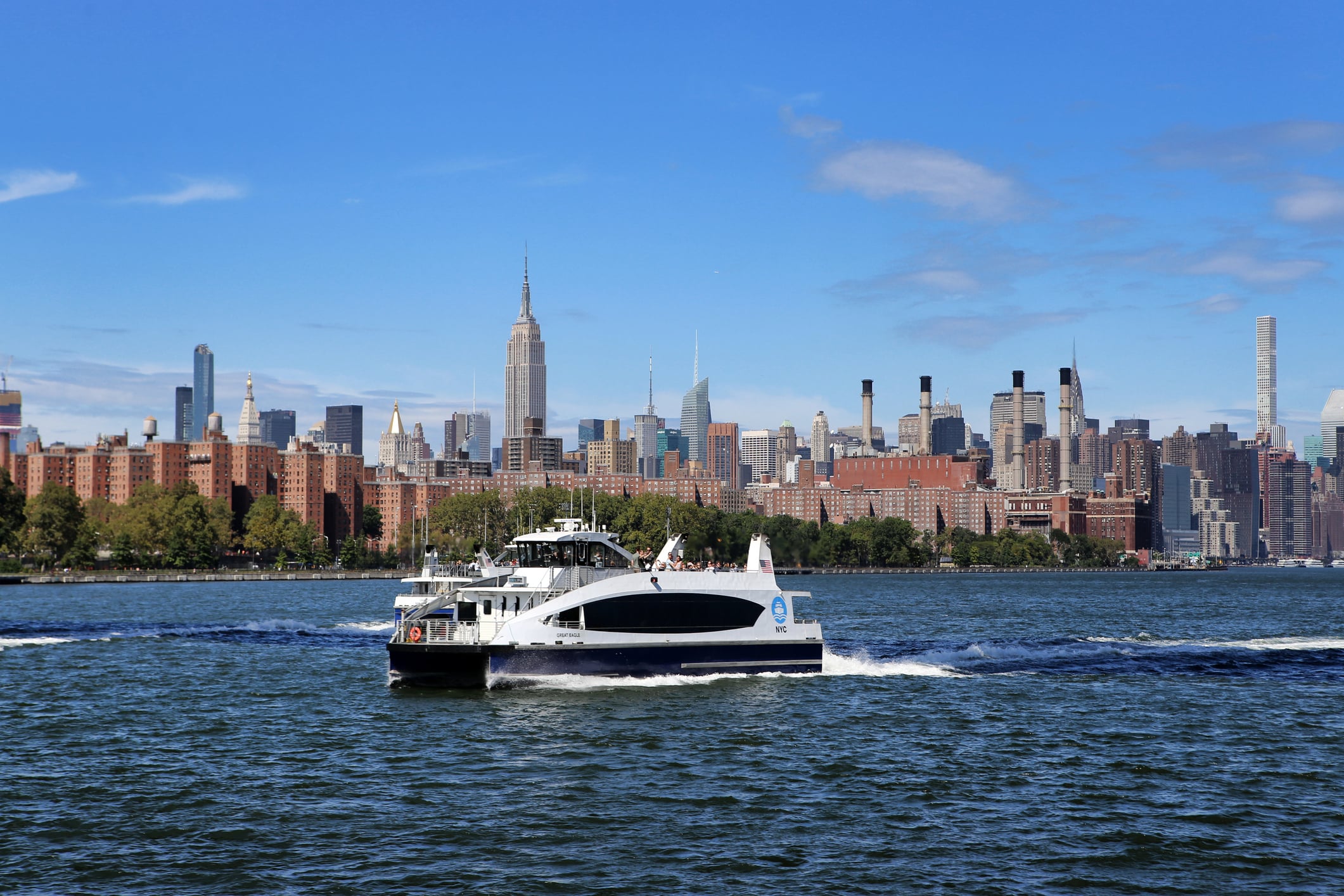 A New York City ferry travels on the East River with the Manhattan skyline, including the Empire State Building, visible in the background on a clear, sunny day.