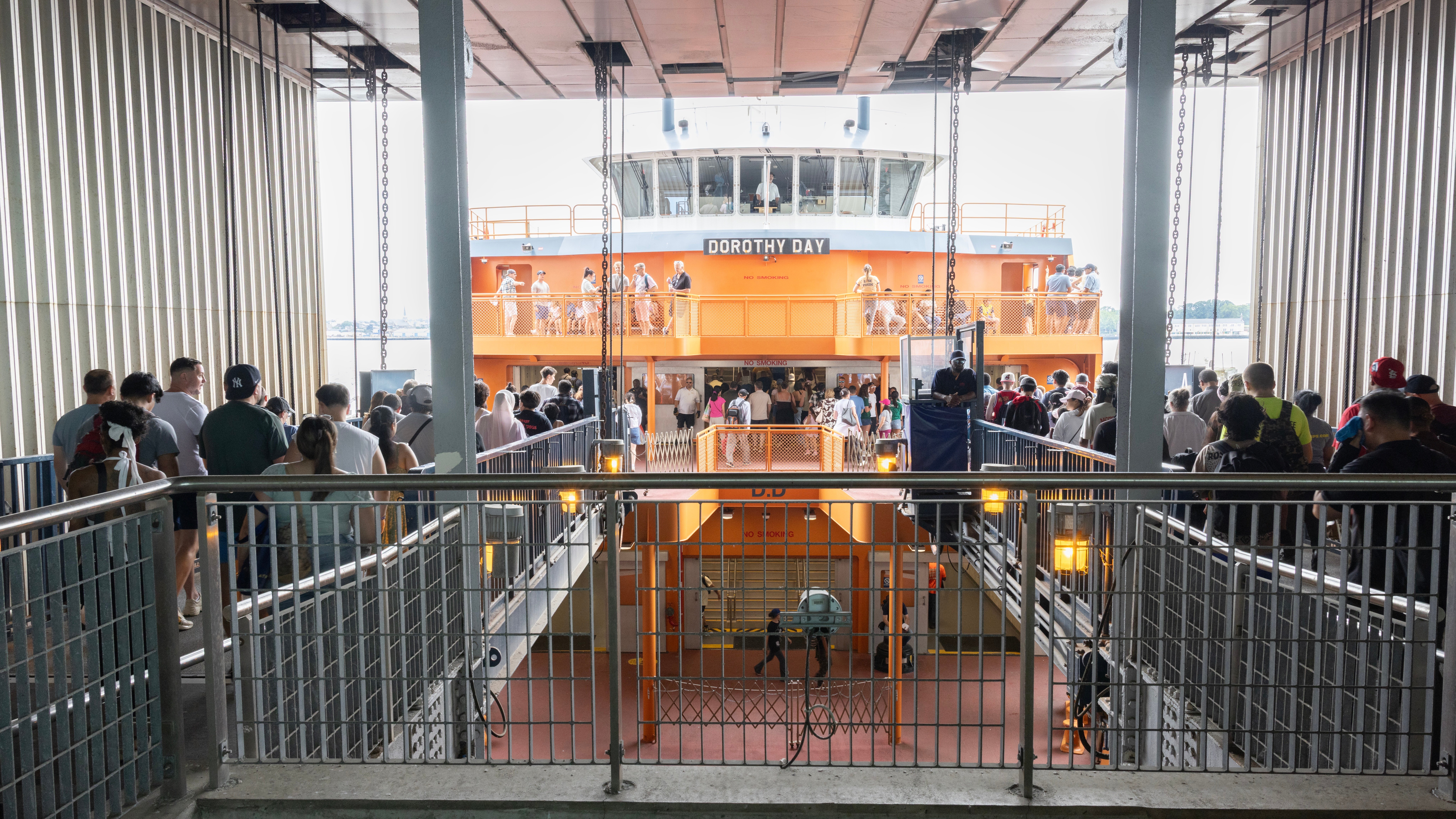 People boarding the orange Staten Island Ferry named Dorothy Day via ramps inside a terminal, with crowds ascending and descending as crew members observe from the ferry.