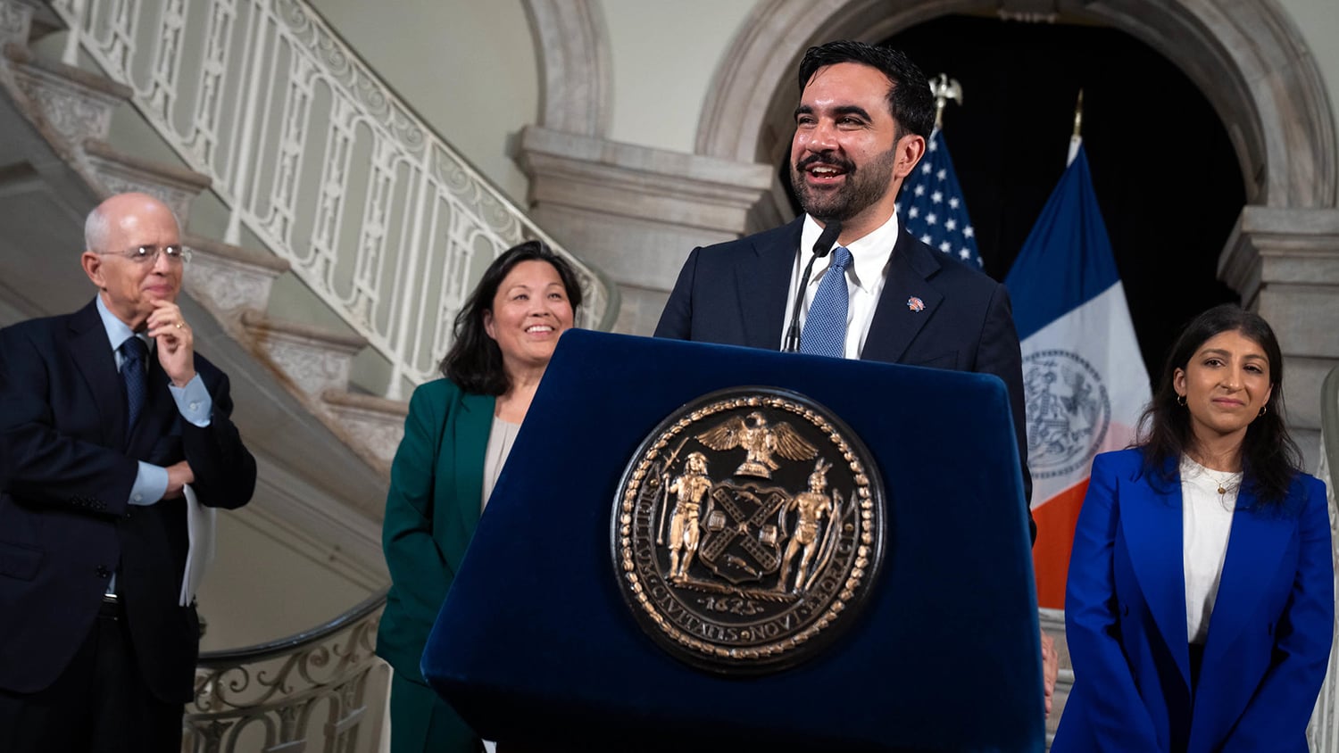 Mayor Mamdani speaks at a podium with the New York City seal, flanked by three people smiling. A staircase and NYC and American flags are in the background.