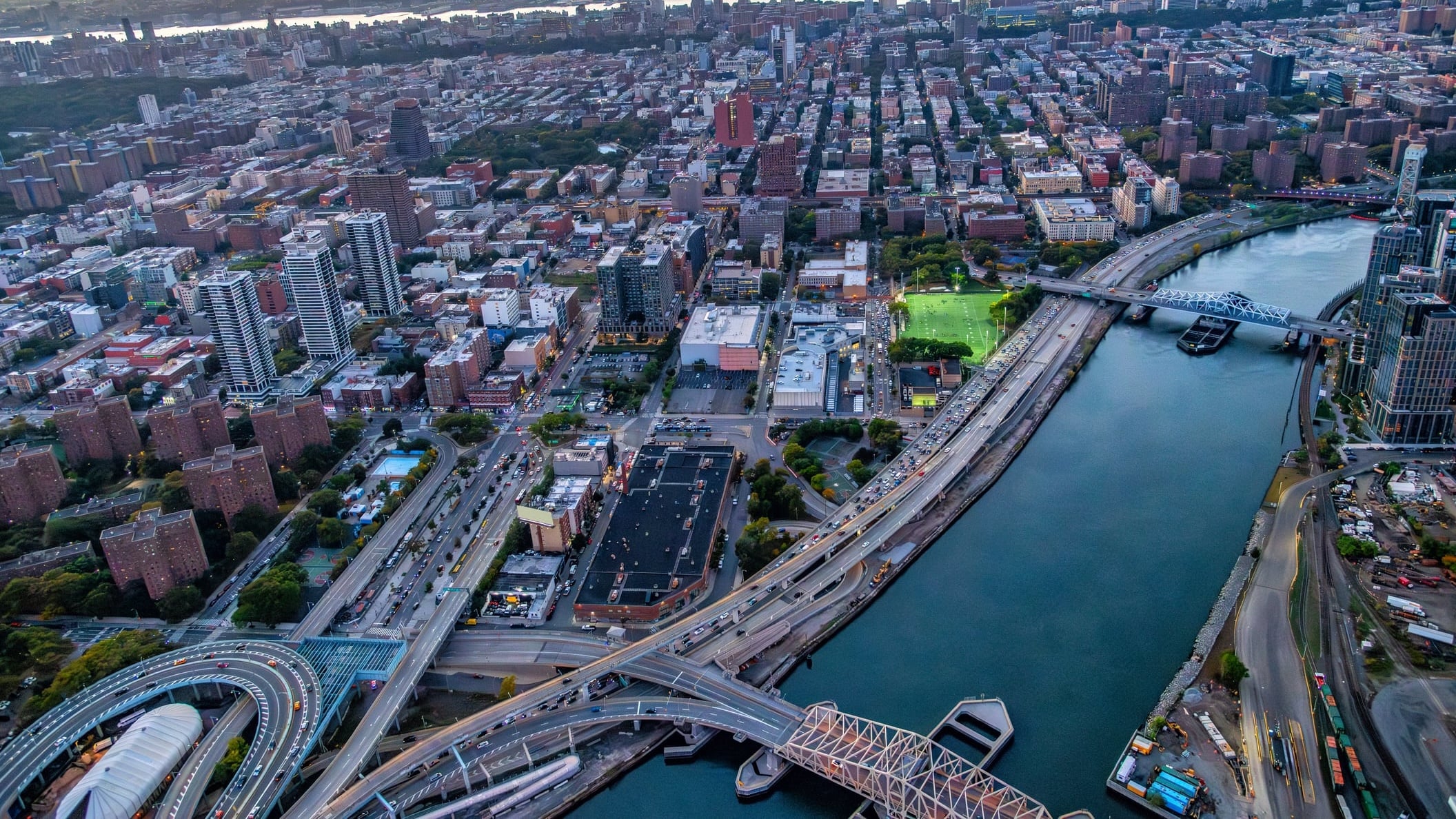 Aerial view of New York City at sunset, showing a river, several bridges, high-rise buildings, busy roads, and a park with green fields.