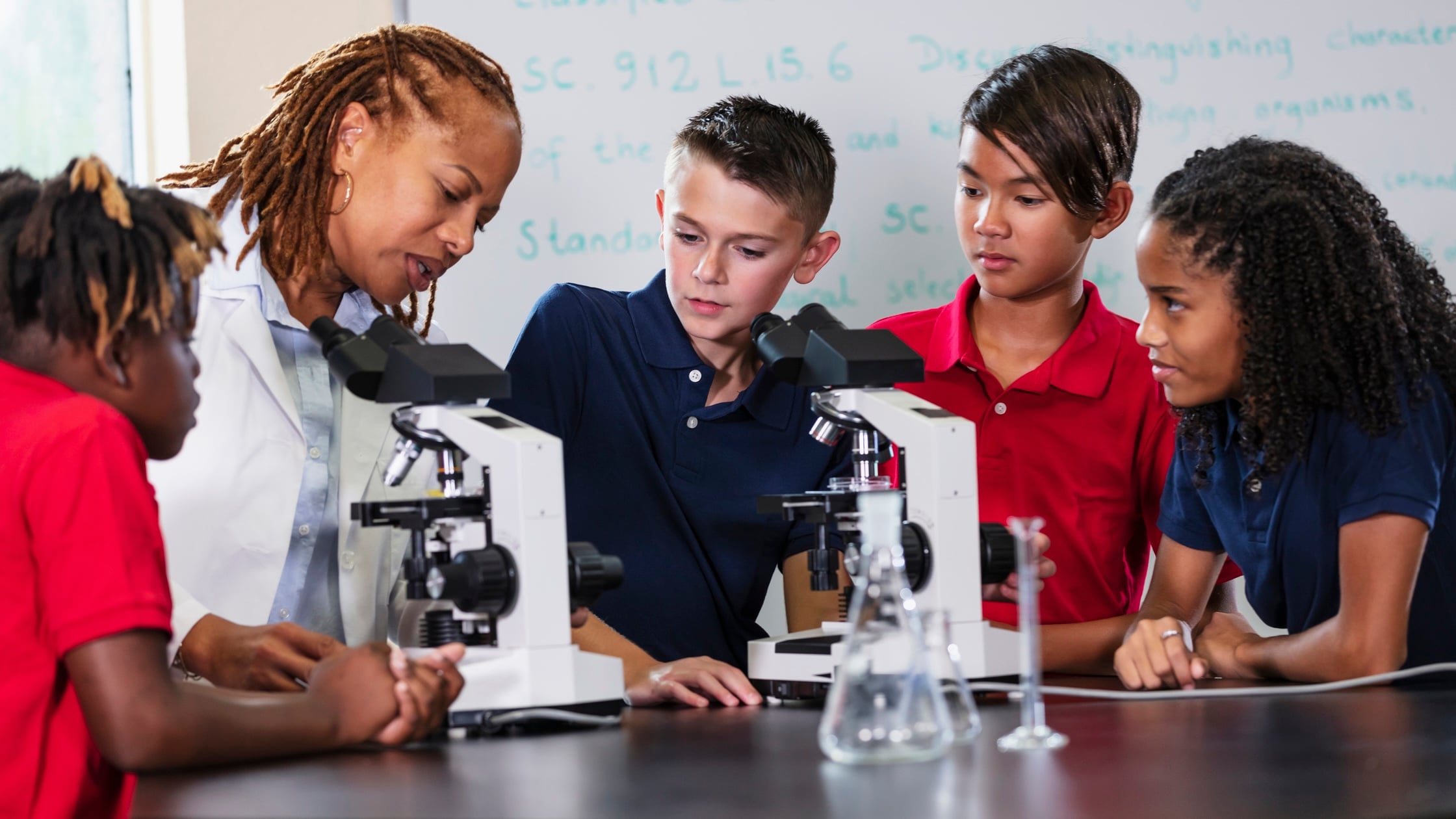 A teacher and four students gather around microscopes in a science classroom, examining slides and discussing their observations. The students look engaged and curious. Laboratory glassware is on the table.