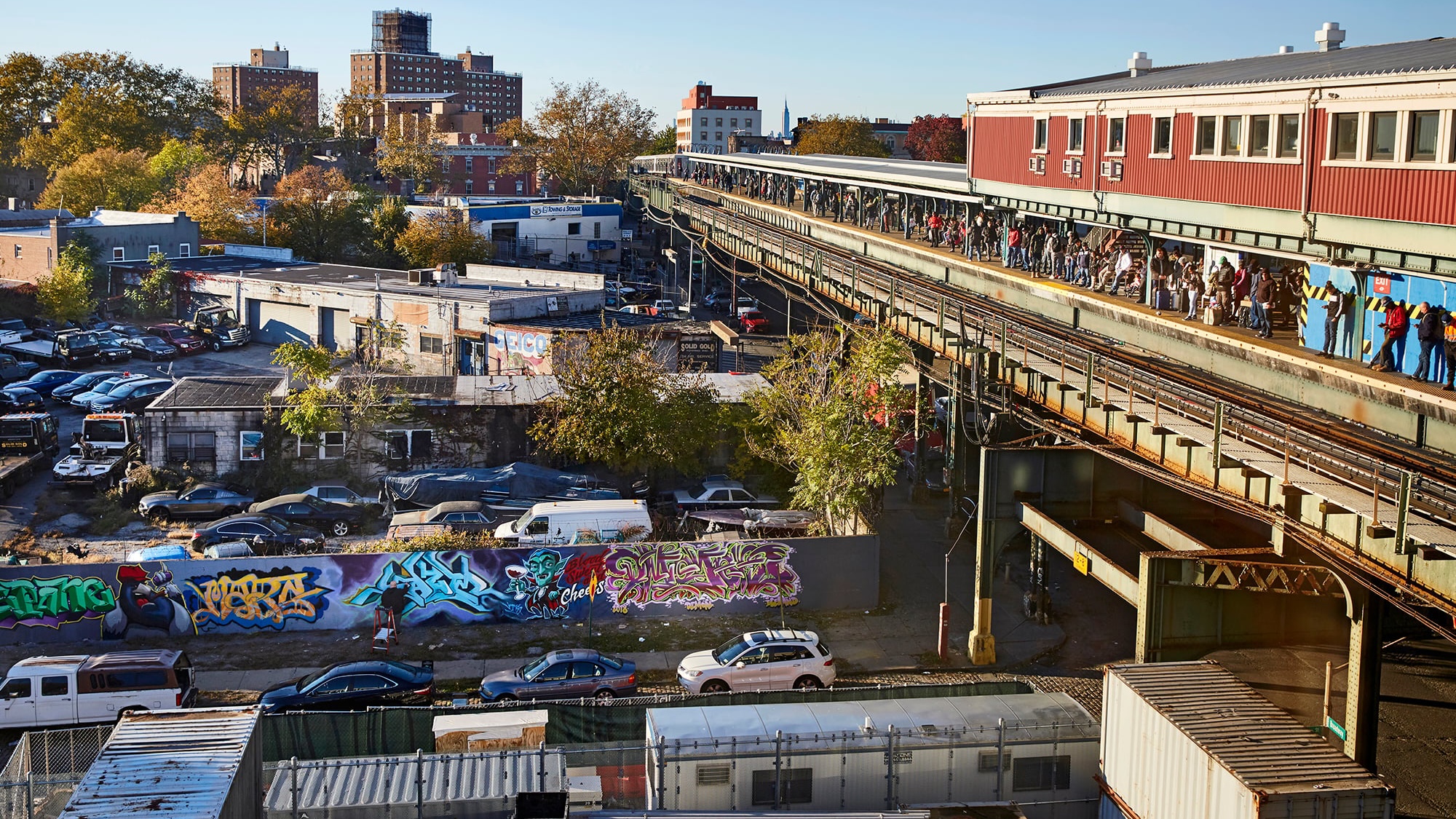 Elevated train platform filled with people runs alongside Broadway Junction, lined with parked cars and colorful graffiti-covered walls. Buildings and trees are visible in the background under a clear blue sky.