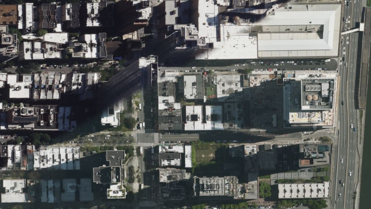 Aerial view of Manhattan blocks with tall buildings casting long shadows over streets, rooftops, and some green spaces; sunlight creates strong contrasts on the urban landscape.