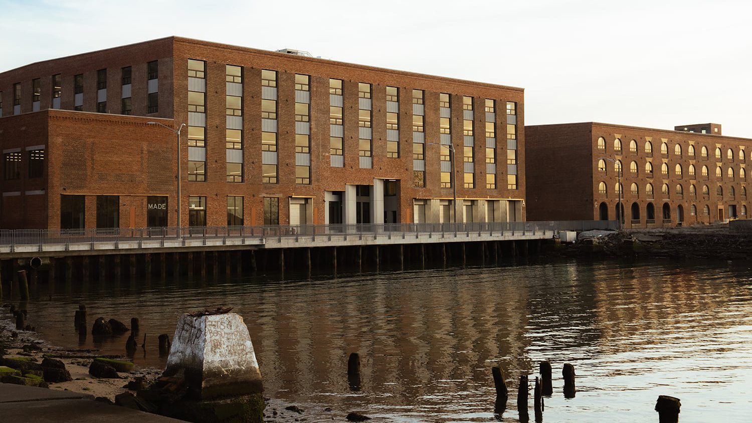 Photos of buildings A and C at MADE Bush Temrinal shows large, rectangular brick buildings with many windows beside a calm waterfront, reflecting in the water. Nearby, weathered wooden posts emerge from the shoreline. The scene is peaceful and bathed in soft light.