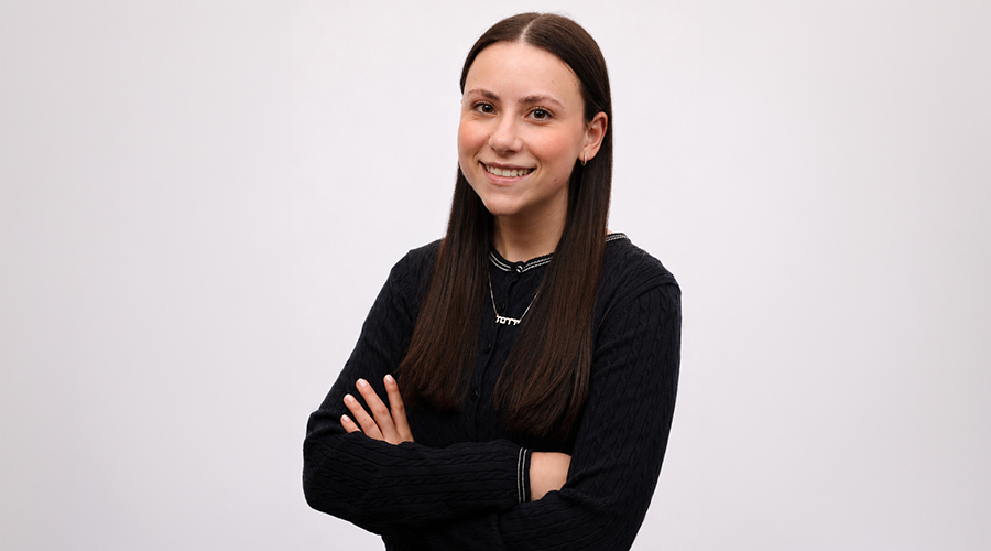 Jordana Stern in long brown hair smiles while standing with her arms crossed. She wears a black sweater and a necklace, and is posed against a plain white background.